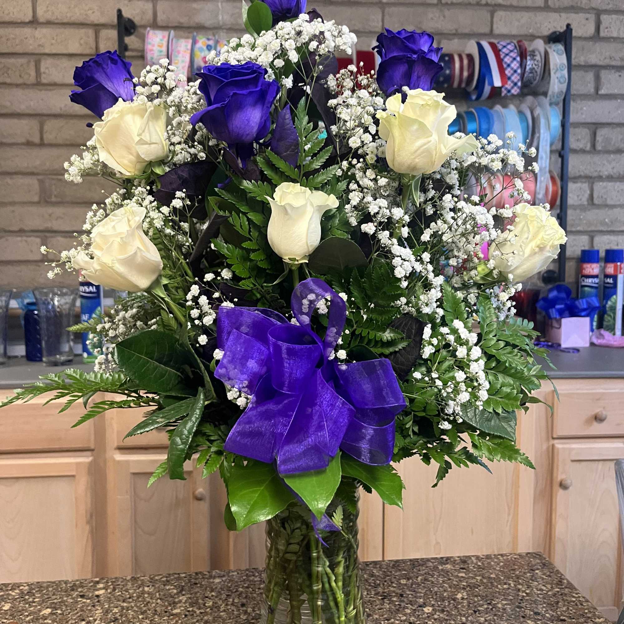 Tall arrangement of purple and white roses with baby's breath in a clear glass vase and purple bow