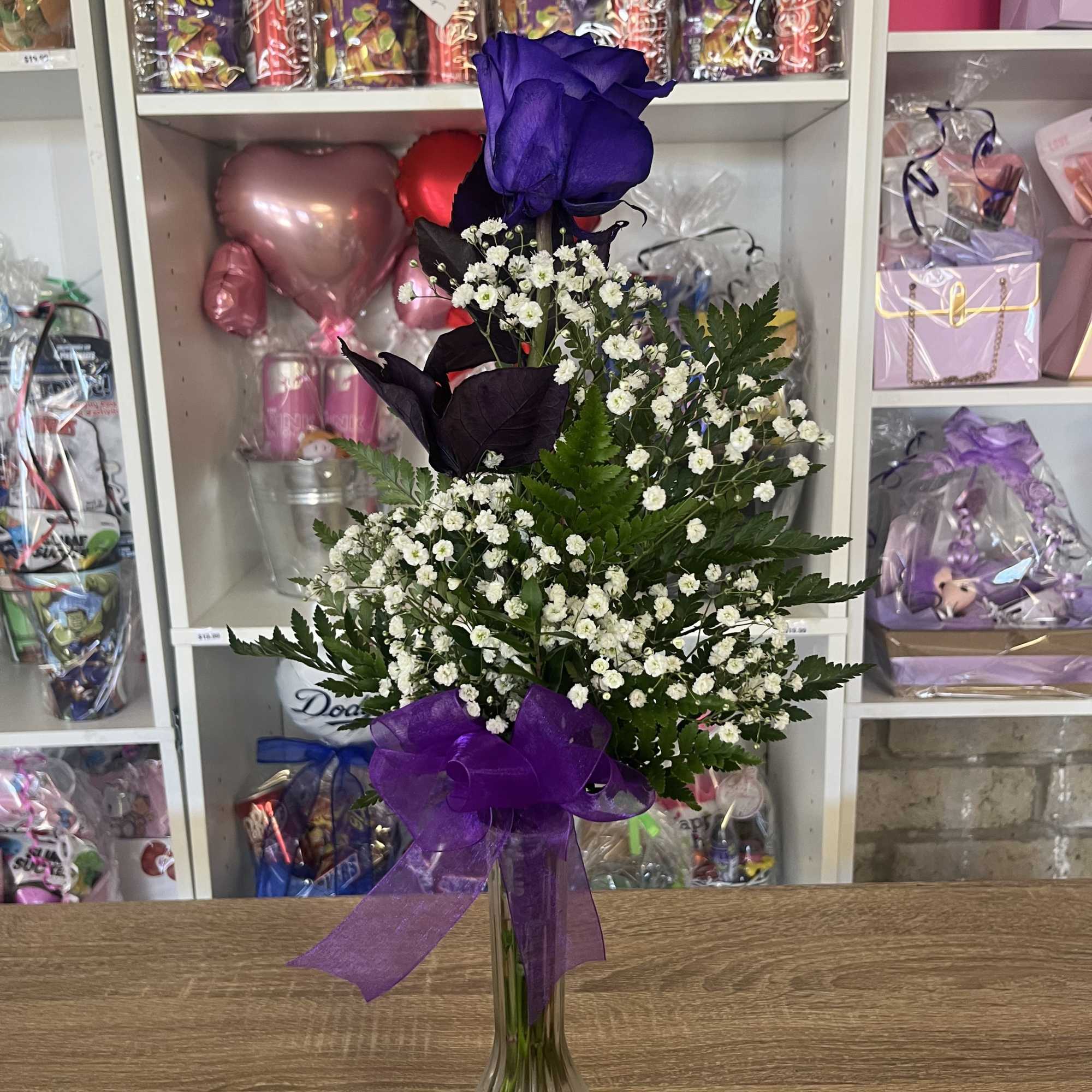 Tall purple rose and white filler flowers in a clear glass vase with a purple bow