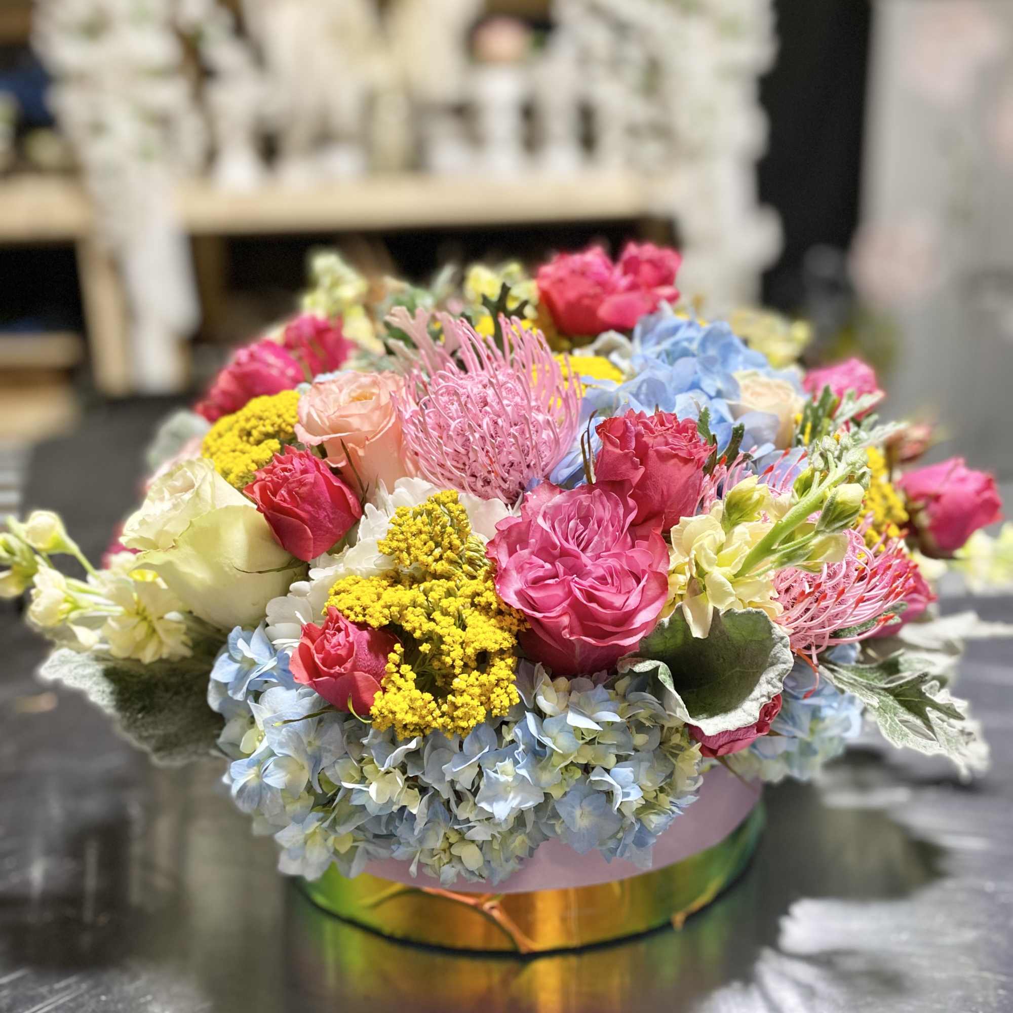 Low round arrangement of pink roses, blue hydrangeas, yellow filler, and pink protea in a glass vase.