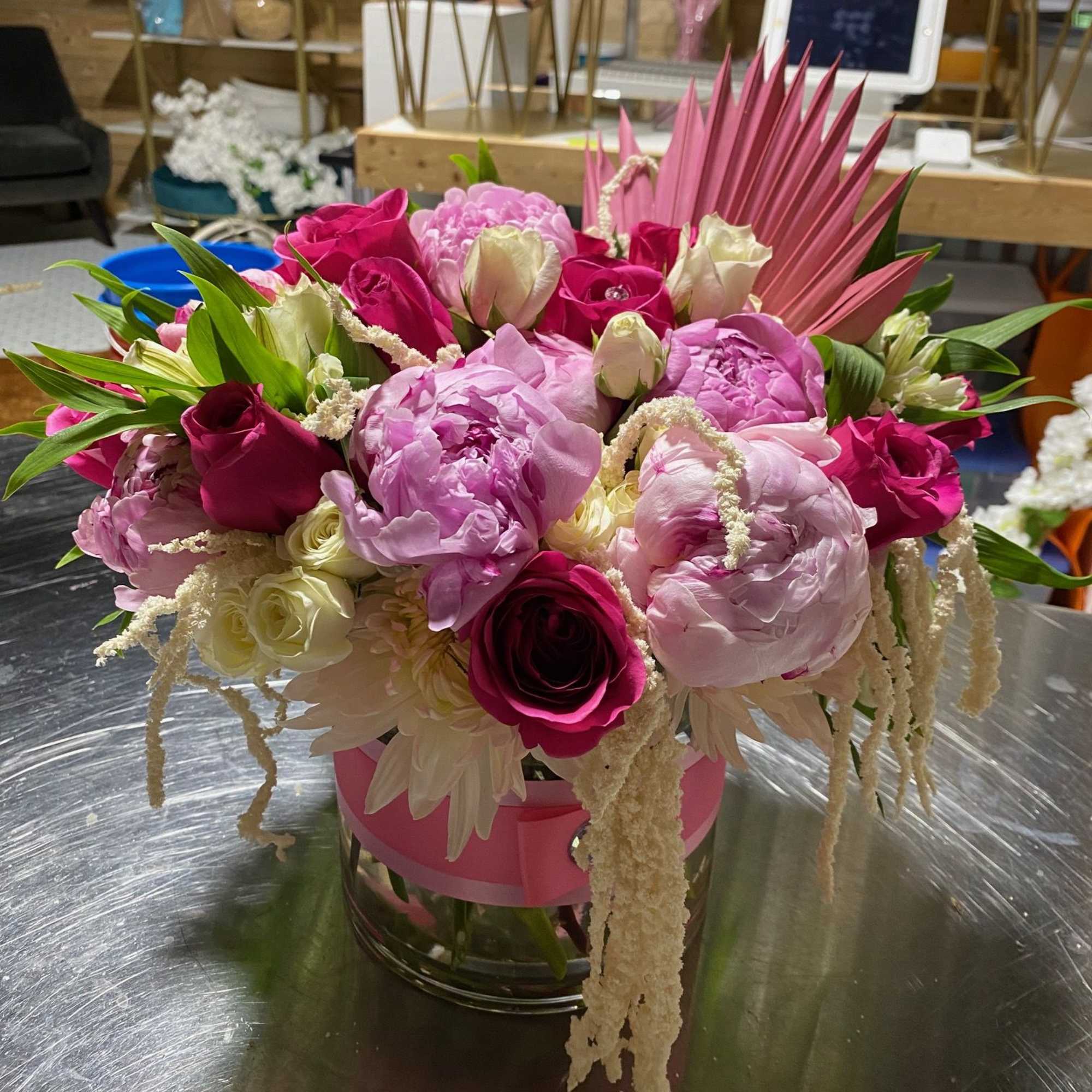 Low arrangement of pink peonies and roses with white blooms in a clear glass vase wrapped with a pink ribbon