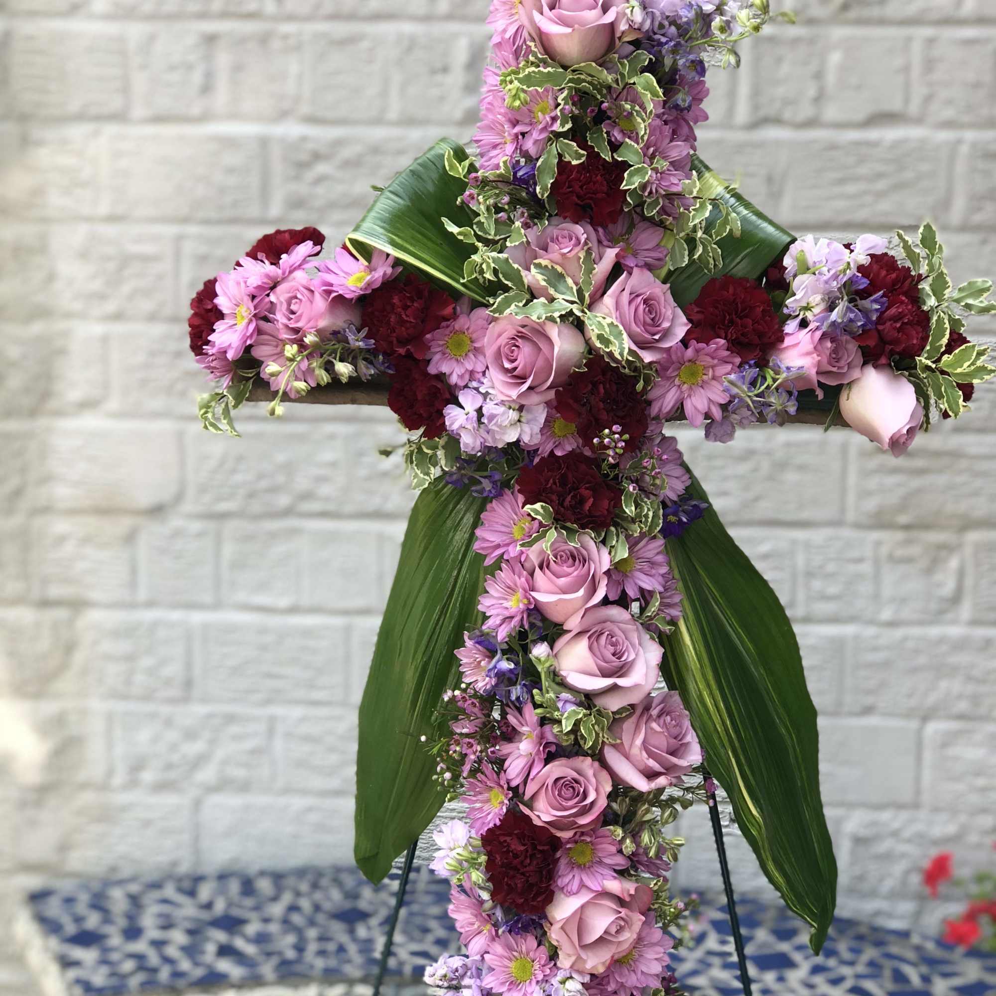 Standing floral cross with pink roses, lavender mums, red carnations, and large green leaves on an easel.