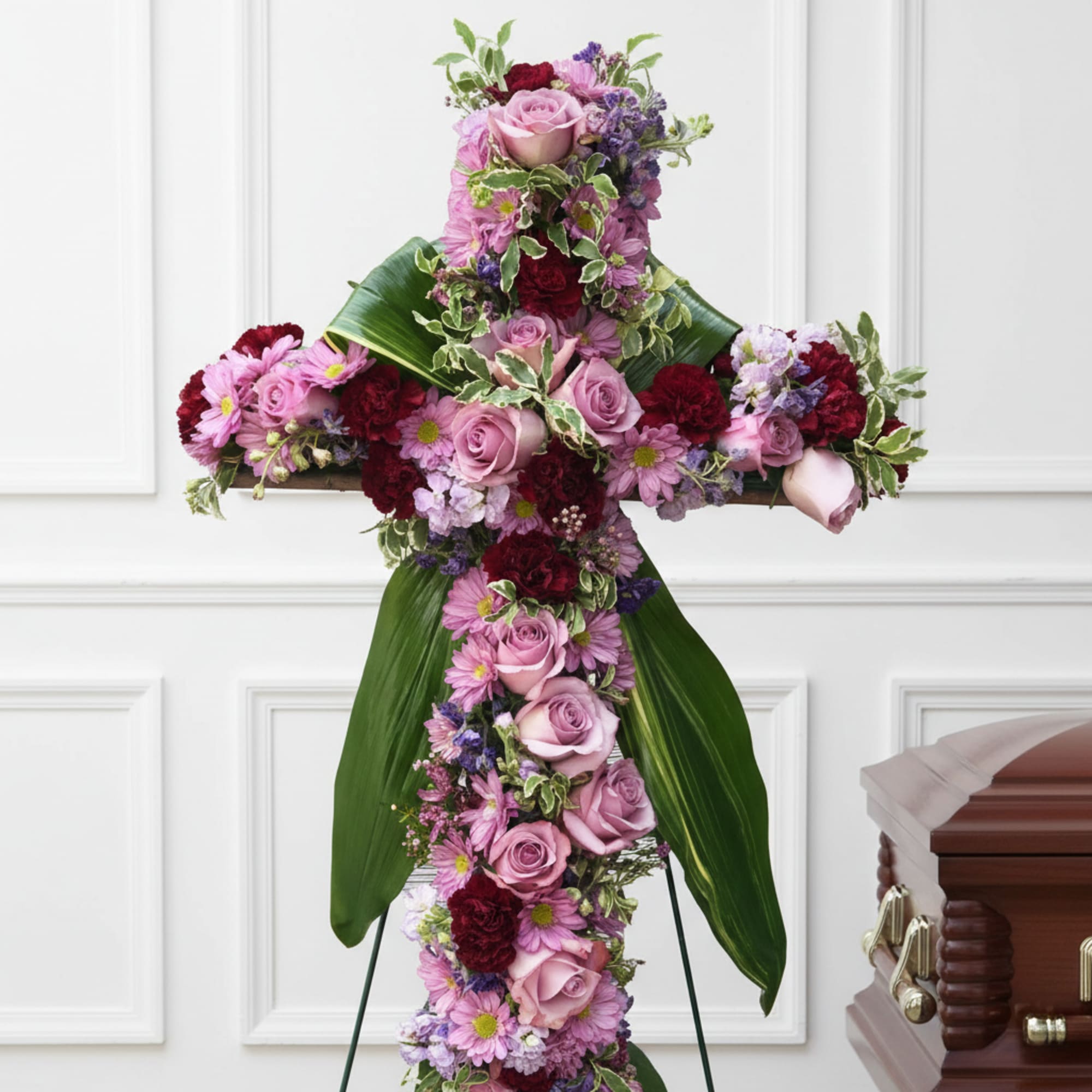 Tall cross-shaped standing spray of pink and burgundy flowers beside a wooden casket.