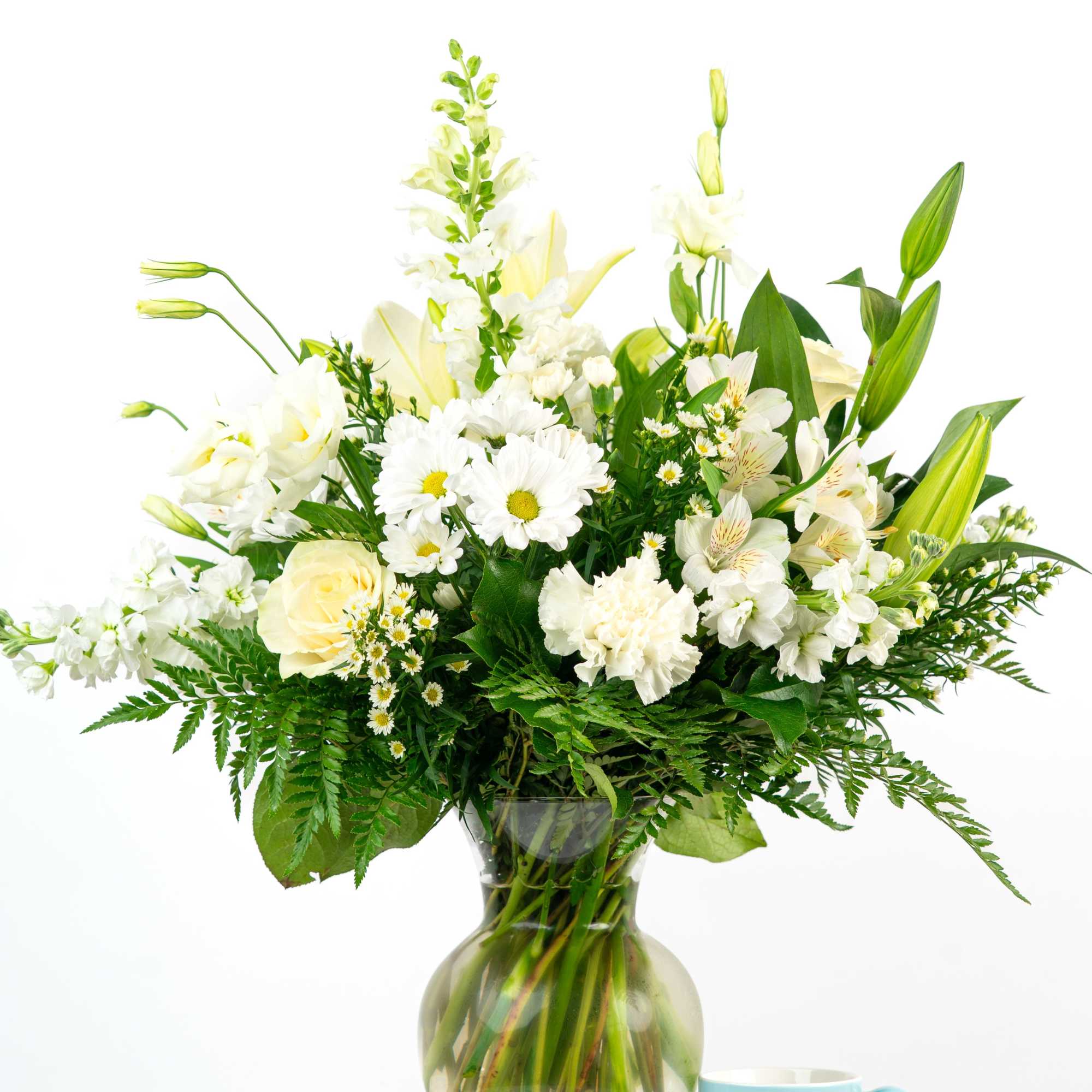 Tall white mixed flower arrangement in a clear glass vase beside a light blue mug