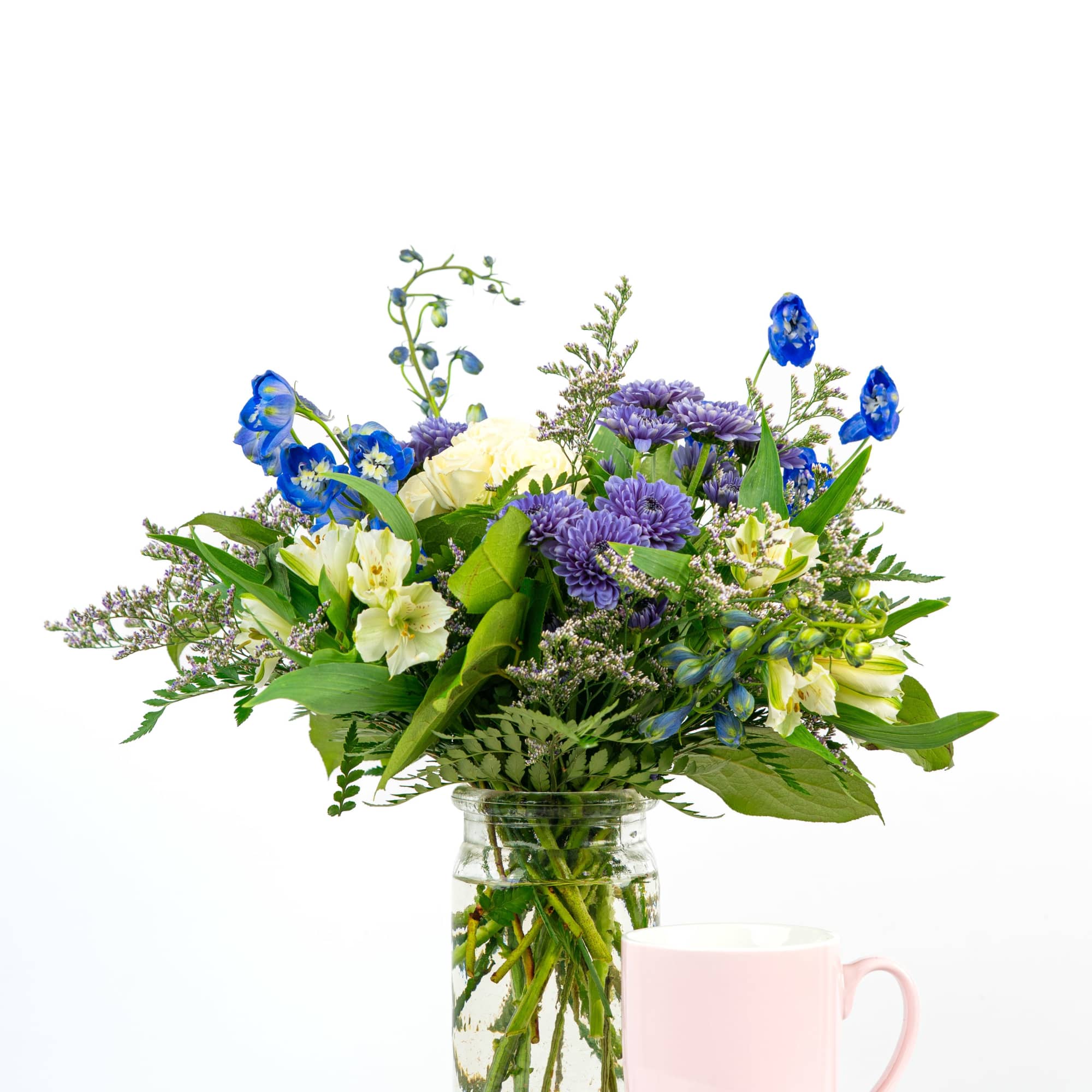 Mixed bouquet of blue, purple, and white flowers in a clear jar vase beside a light pink mug.