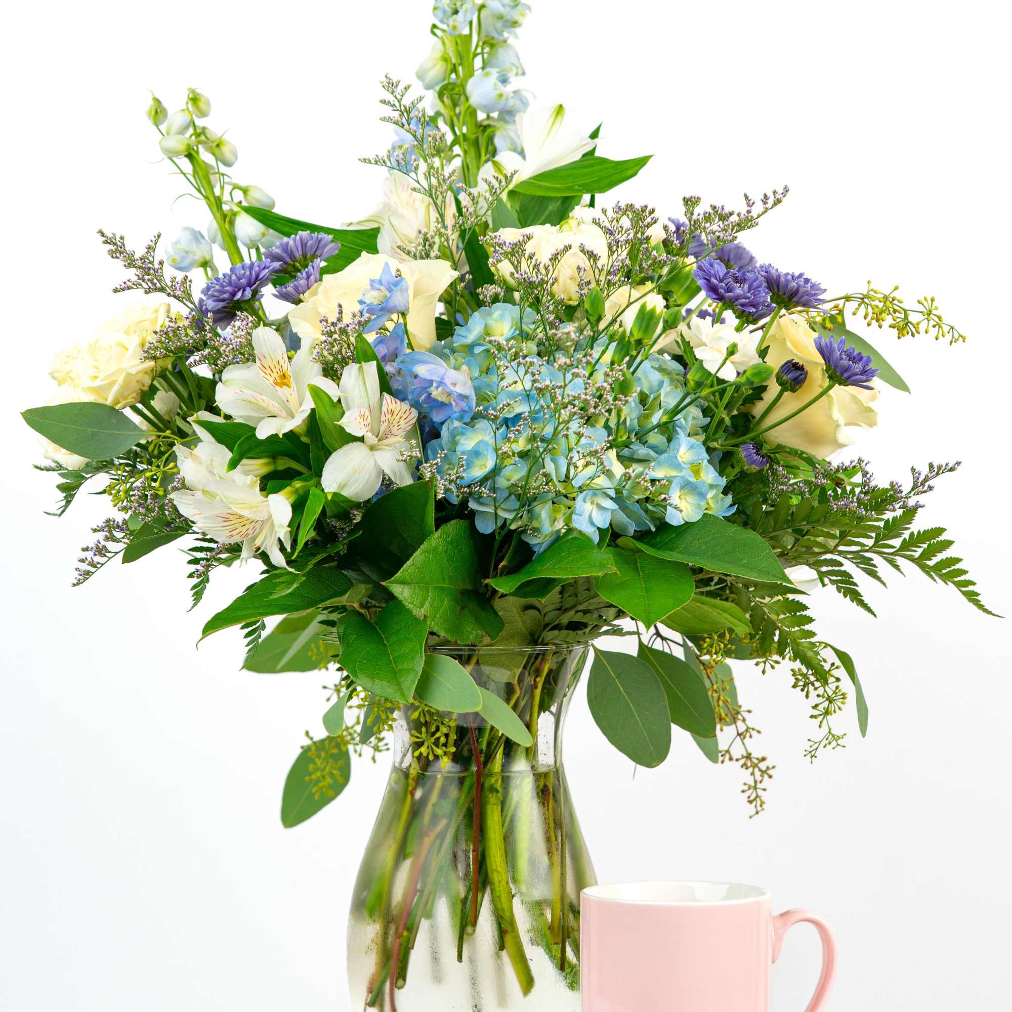 Mixed bouquet of white and blue flowers with purple accents in a clear vase beside a pink mug