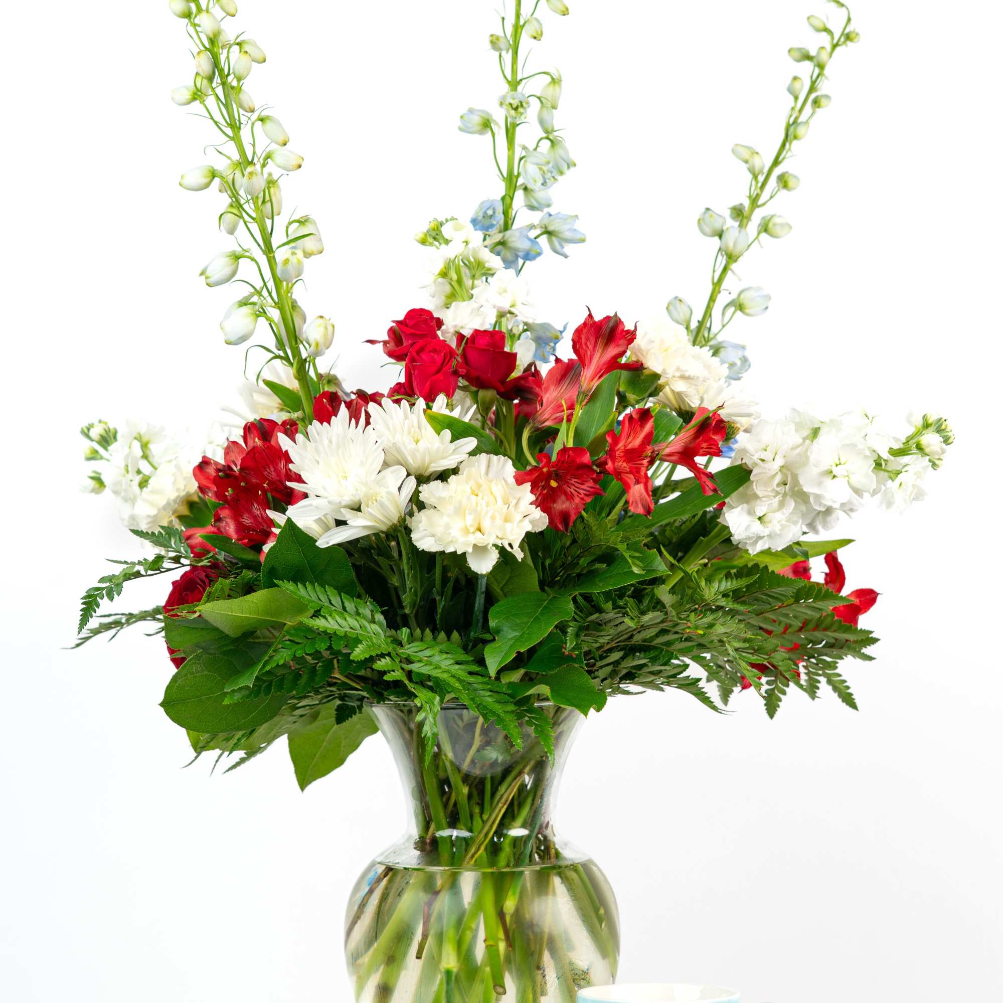 Tall red, white, and blue mixed flower arrangement in a clear glass vase beside a light blue mug