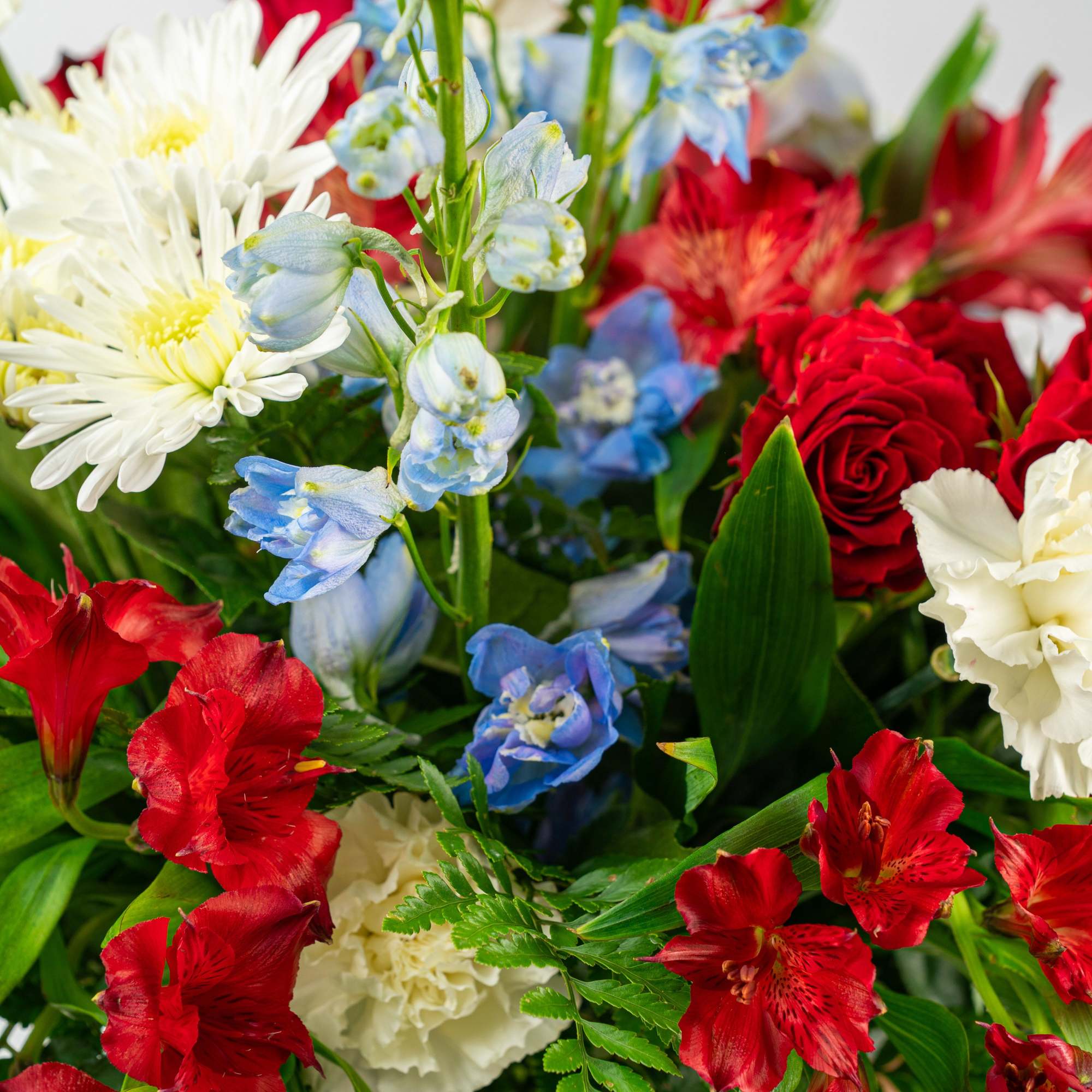 Red, white, and blue mixed flower arrangement with roses, delphinium, mums, and carnations in a clear glass vase