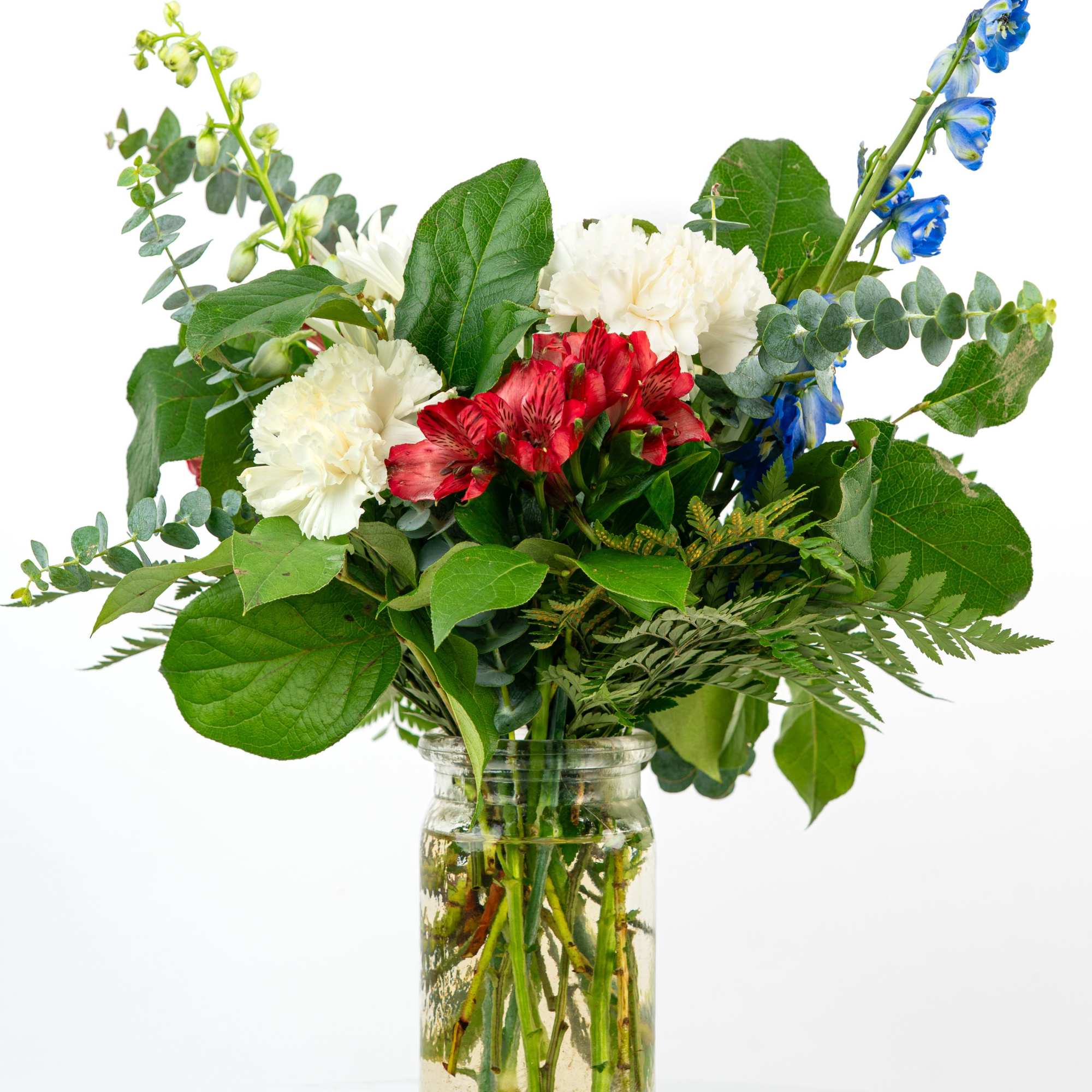 Red, white, and blue mixed flower arrangement with lush foliage in a clear glass jar vase