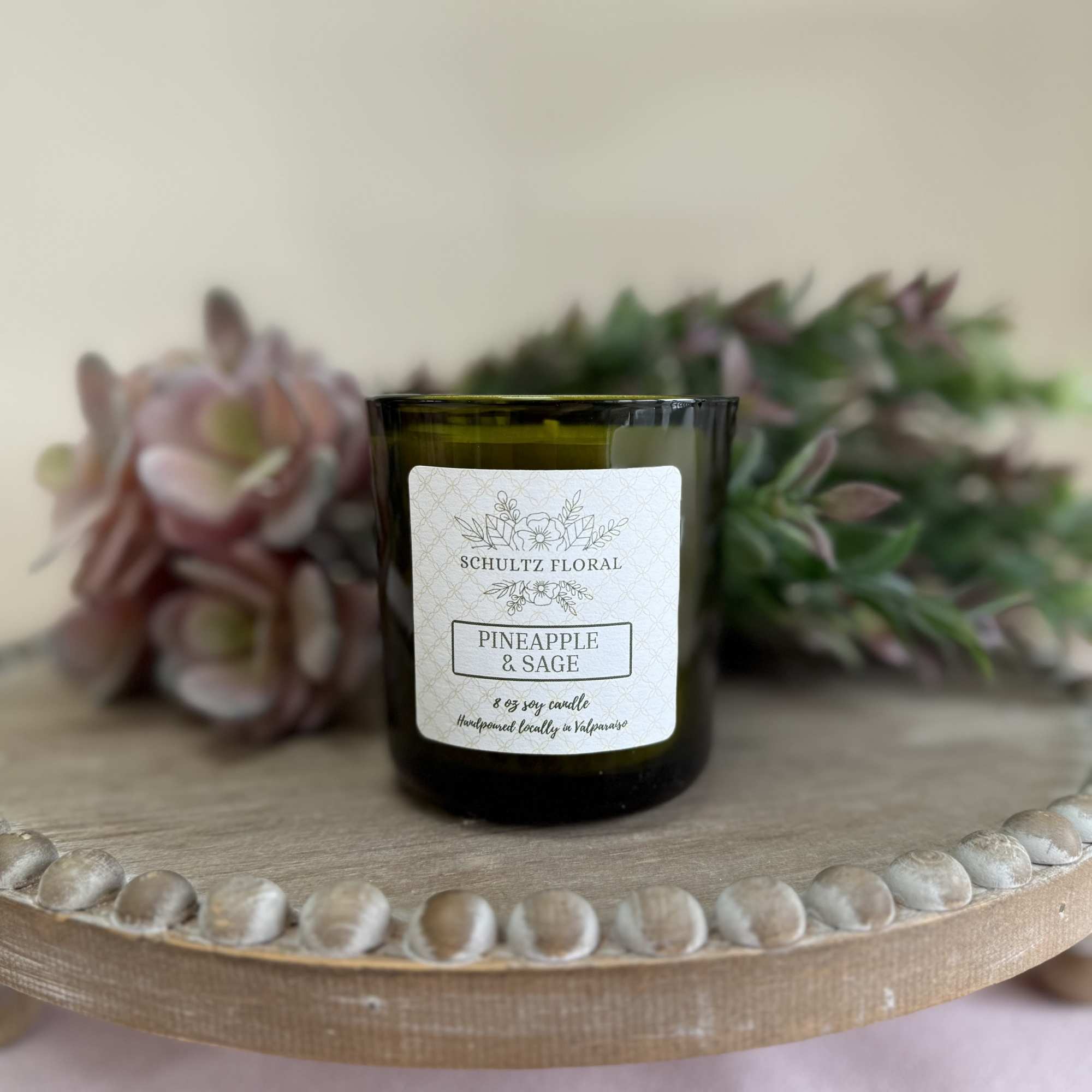 Green glass pineapple and sage candle on a wooden stand with soft pink and green florals behind.