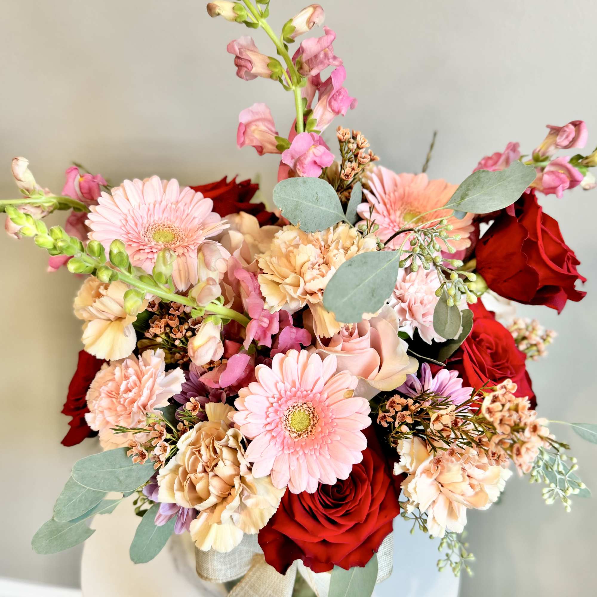 Mixed arrangement of red roses, pink gerbera daisies, carnations, and snapdragons in a glass vase with ribbon.