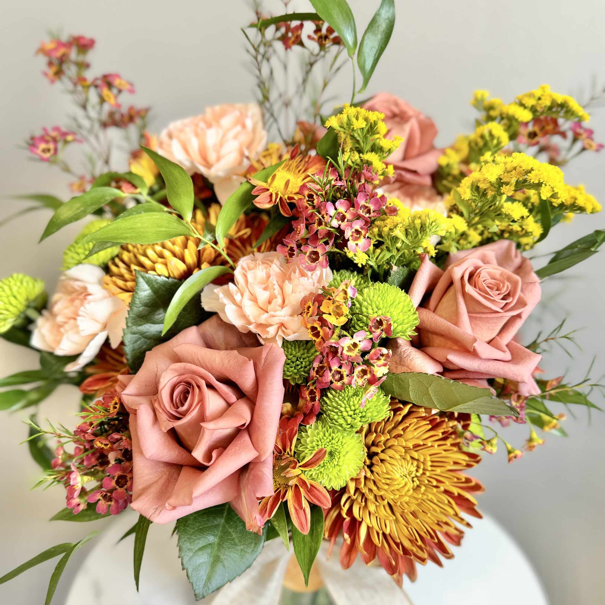 Peach roses, carnations, yellow mums, and green button poms arranged in a clear glass vase with a bow