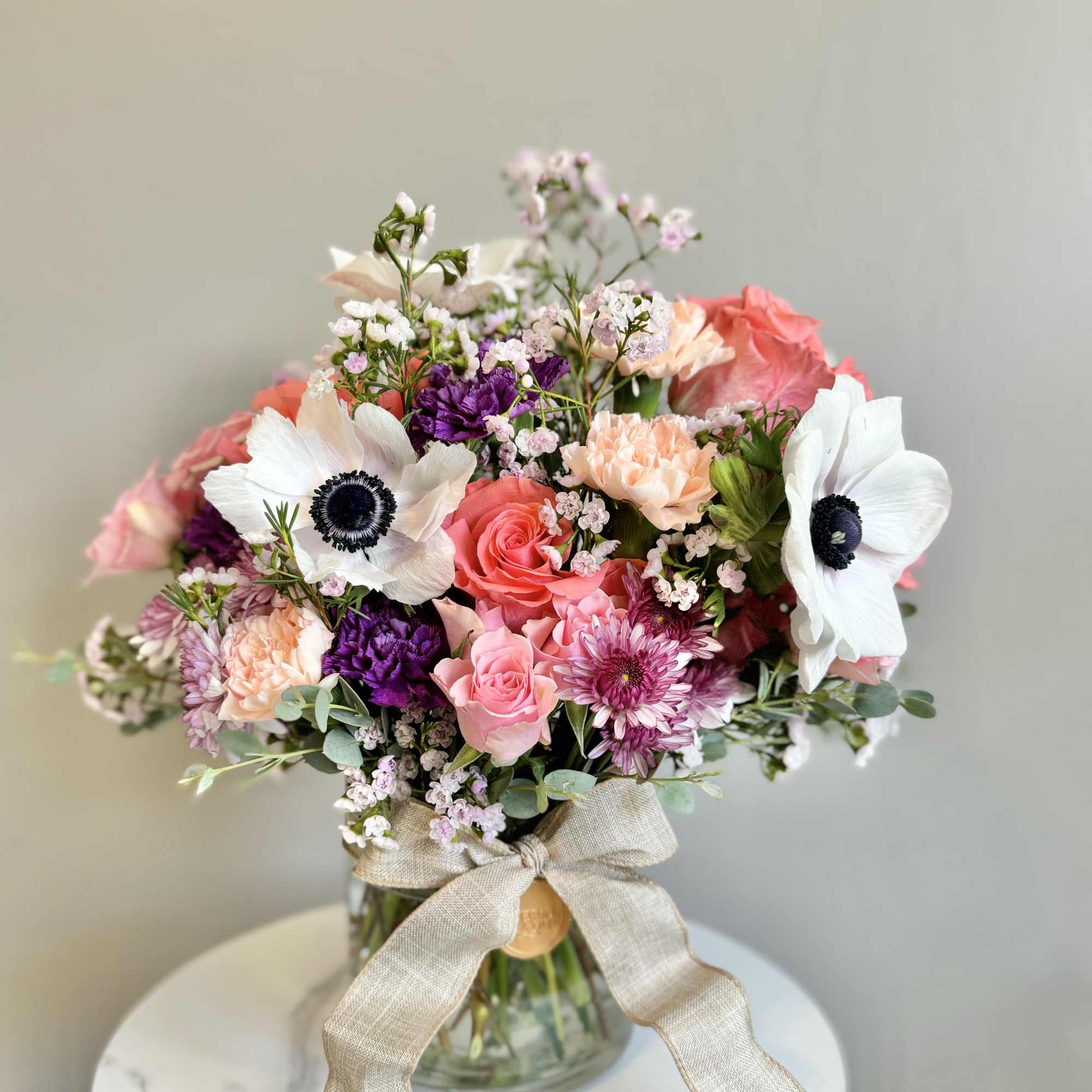 Mixed bouquet of pink roses, white anemones, and purple blooms in a glass vase with a linen bow.