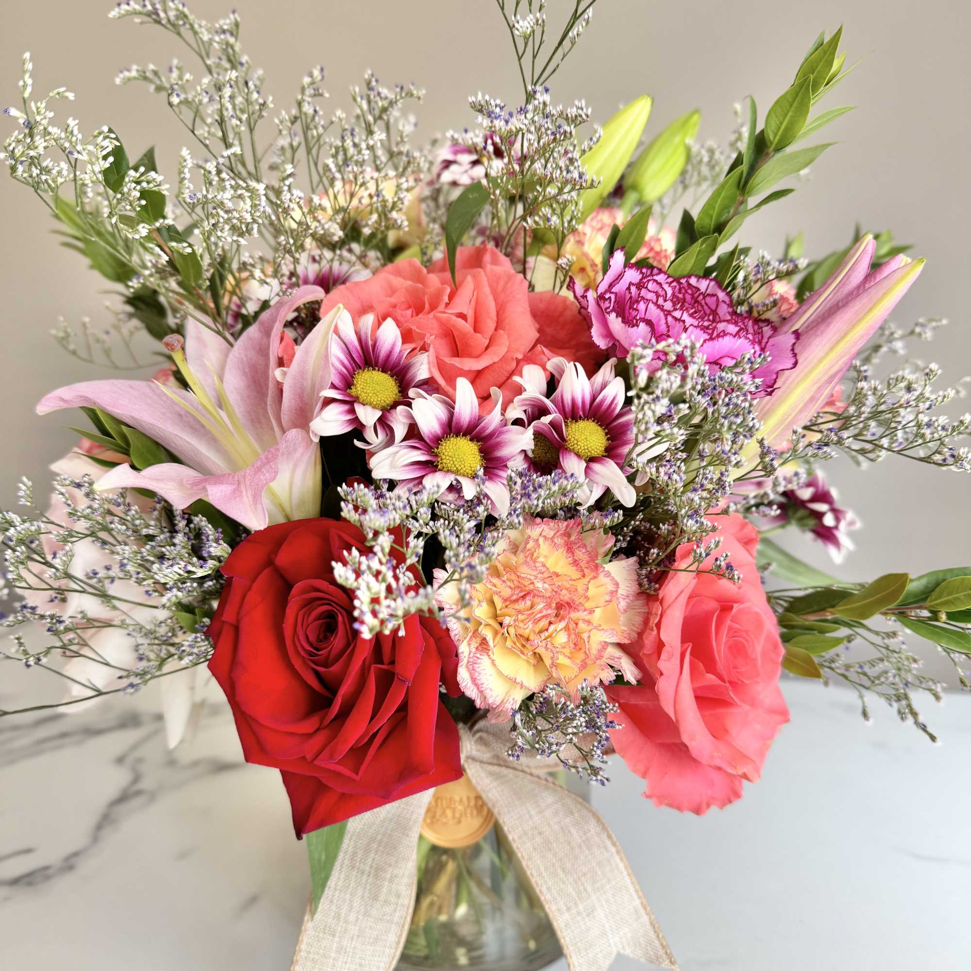 Mixed arrangement of pink lilies, red and coral roses, carnations, and daisies in a glass vase with a bow