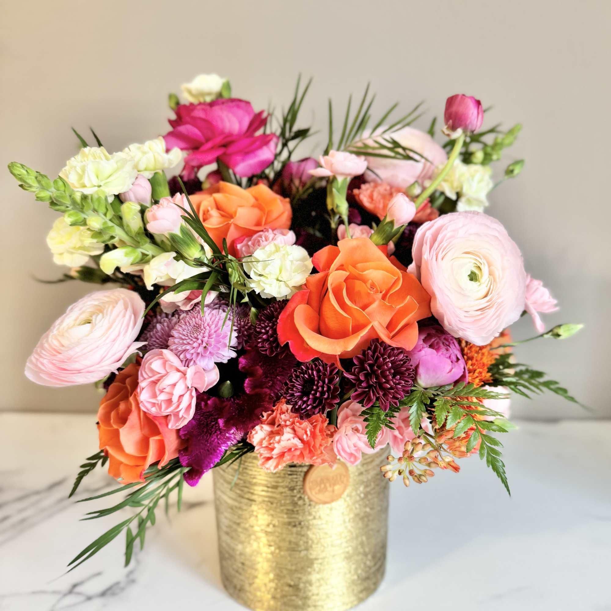 Bright arrangement of orange roses and pink blooms in a gold cylinder vase on a marble surface