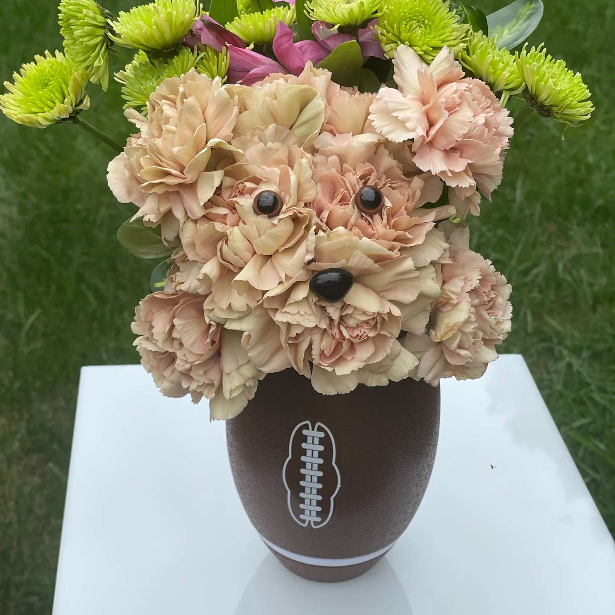 Dog-shaped peach carnation arrangement with green mums in a brown football vase on a white stand.