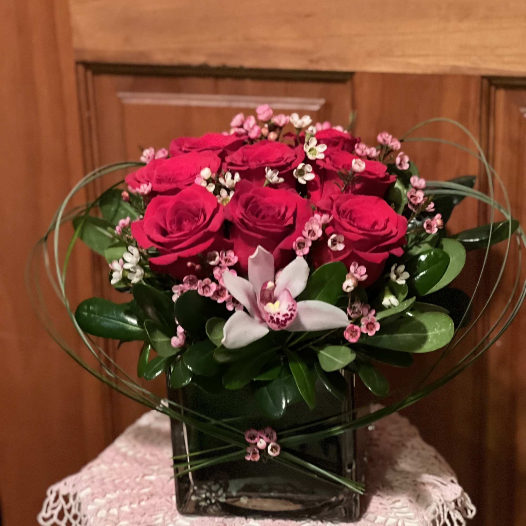 Low arrangement of red roses, a pink orchid, and tiny pink blooms in a black cube vase with looping greenery.
