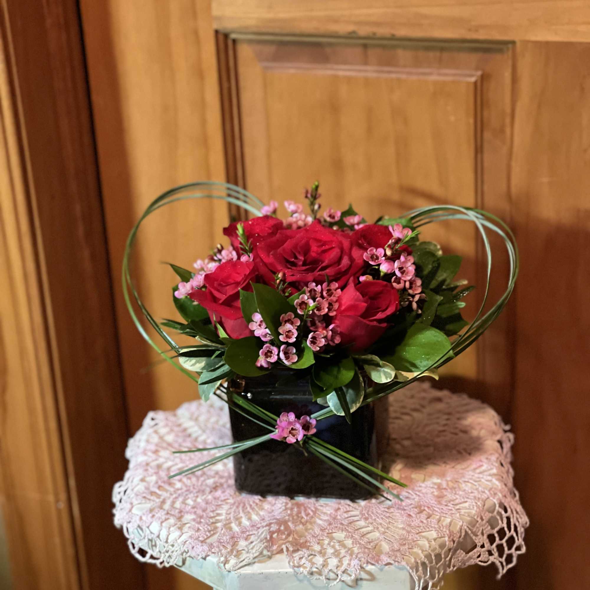 Compact arrangement of red roses and pink blossoms in a black cube vase with heart-shaped loops of grass.