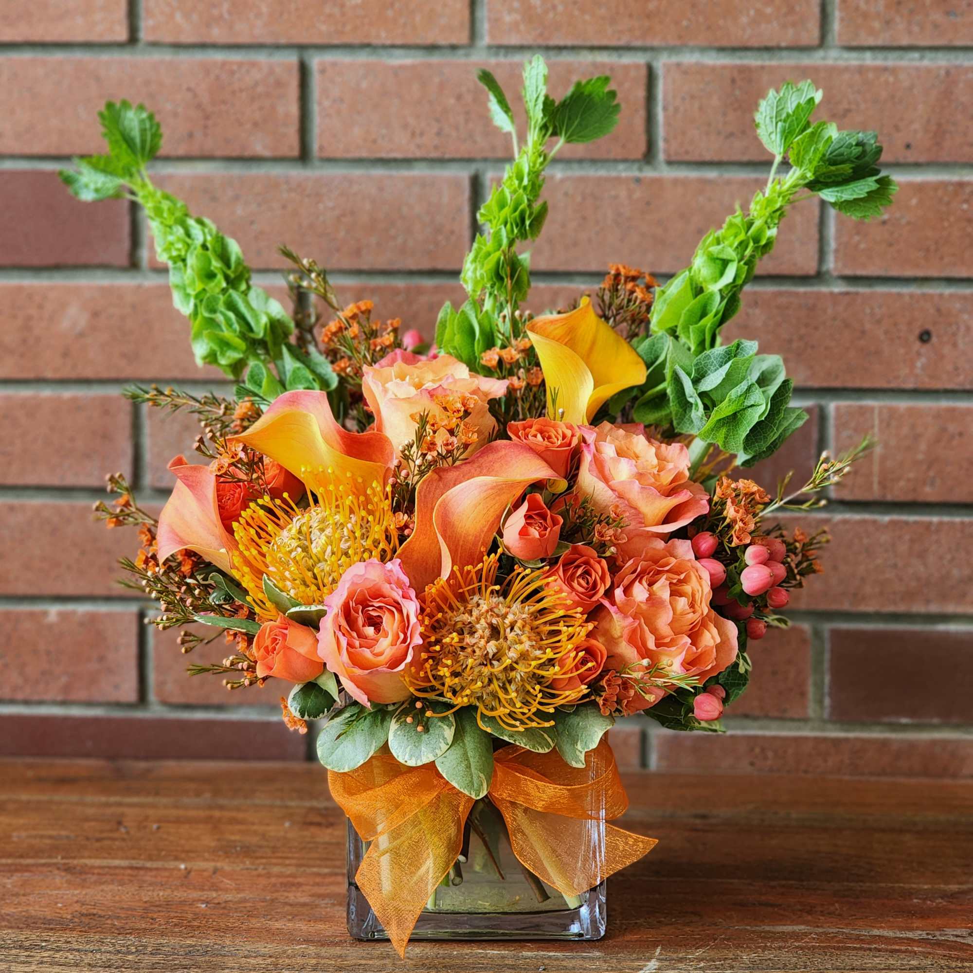 Arrangement of orange roses, calla lilies, protea and Bells of Ireland in a glass cube vase with an orange bow