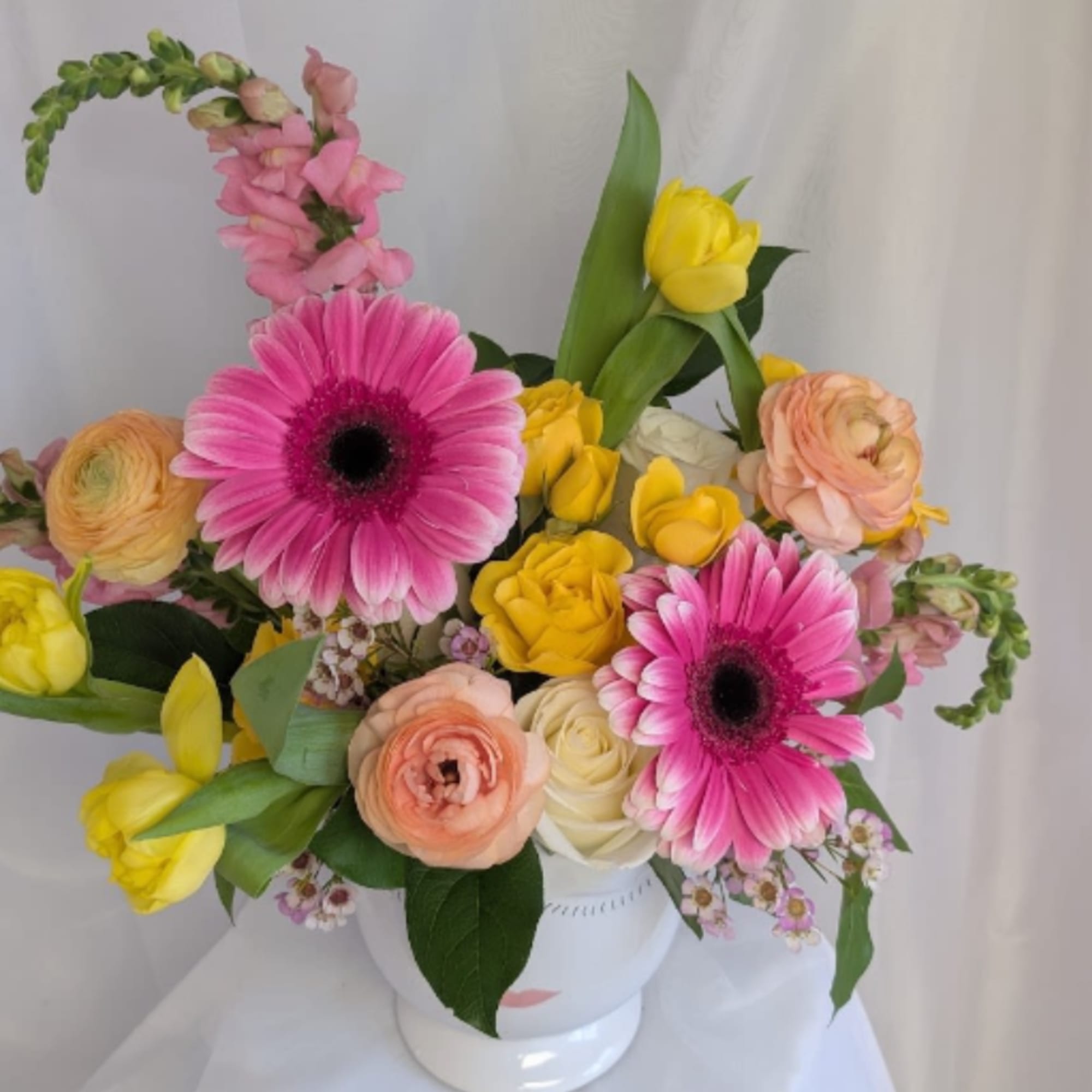 Bright arrangement of pink gerbera daisies, yellow tulips, and roses in a white face vase.