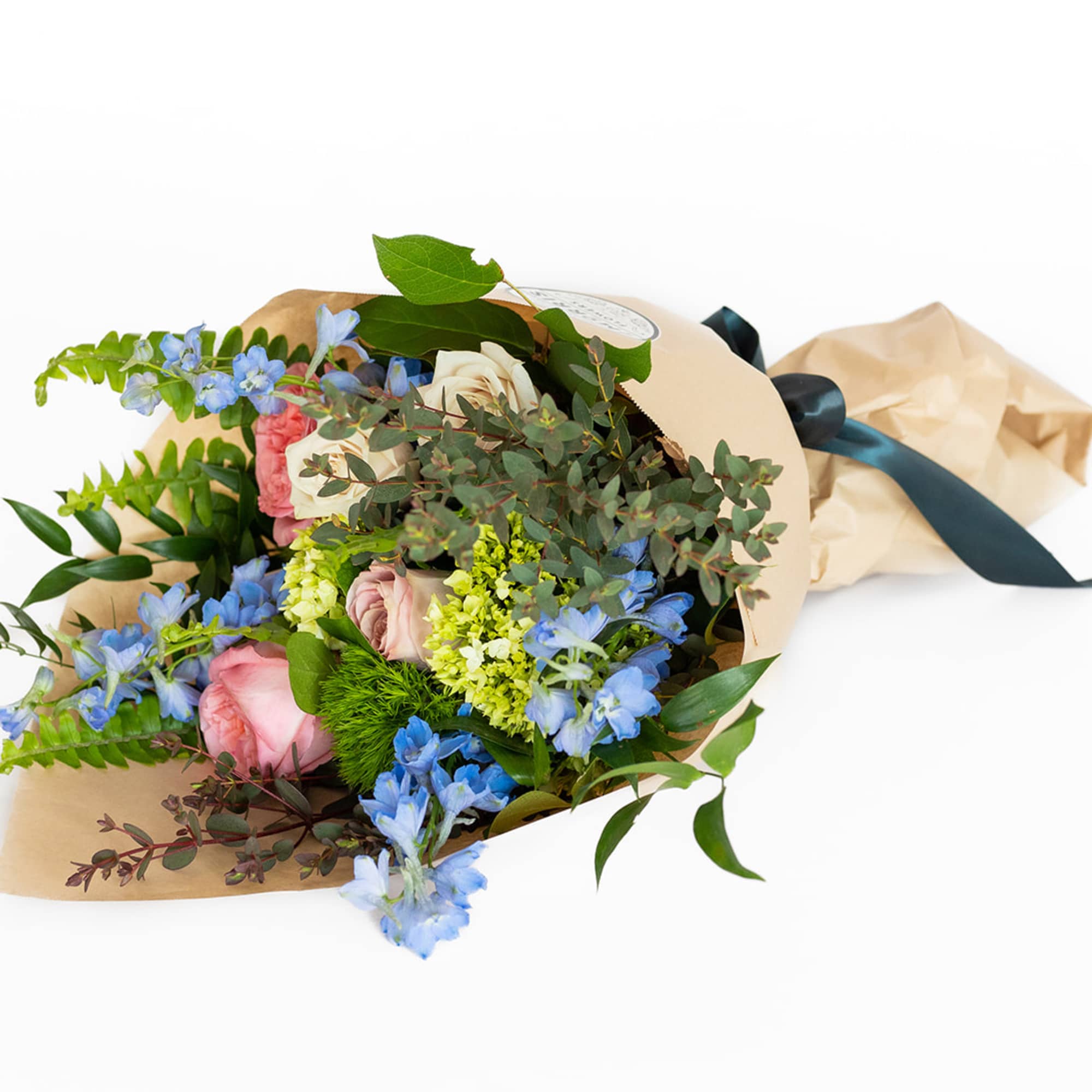 Wrapped bouquet of pink and cream roses with blue accent flowers in kraft paper tied with a dark ribbon