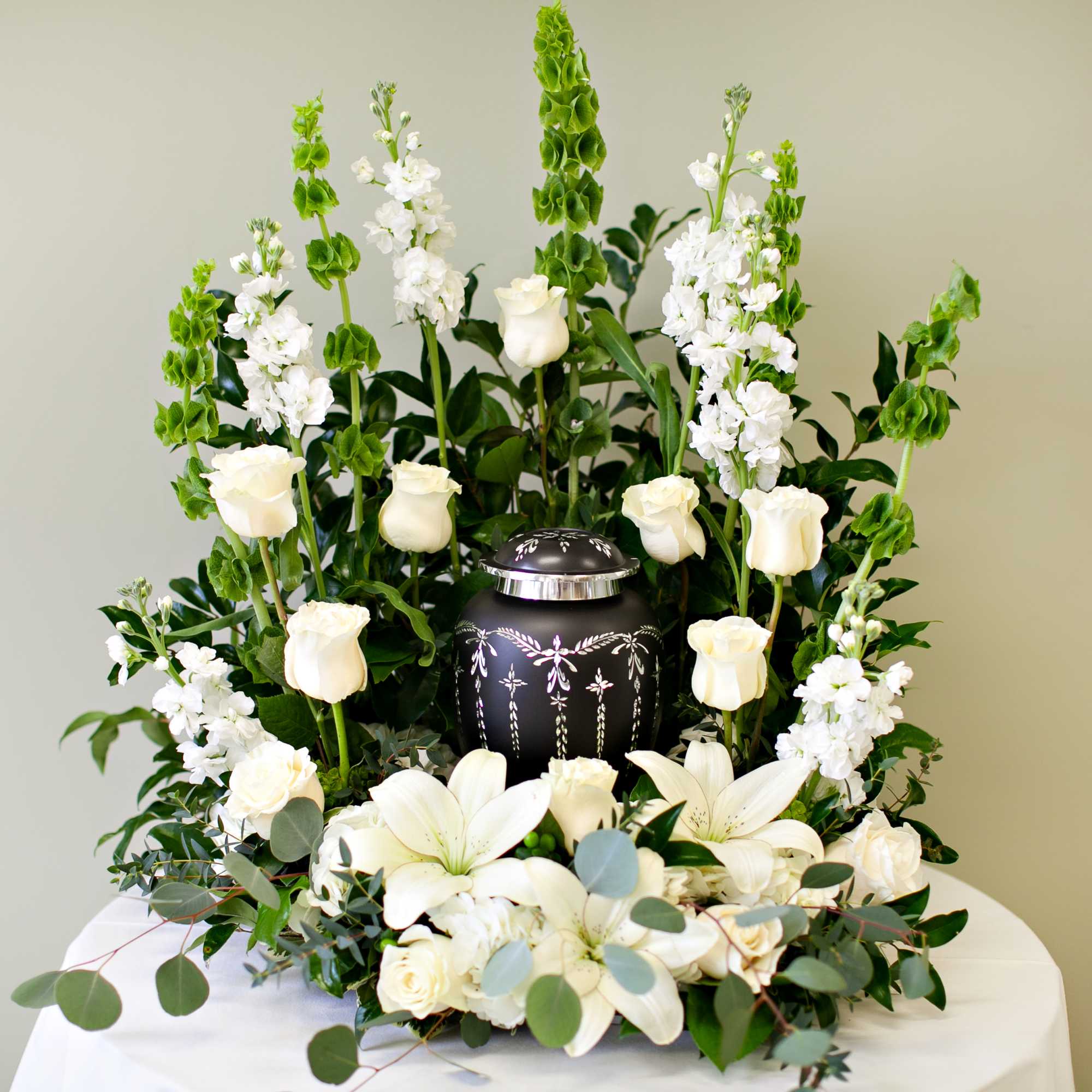 White roses and lilies surrounding a decorative urn in a tall white sympathy arrangement