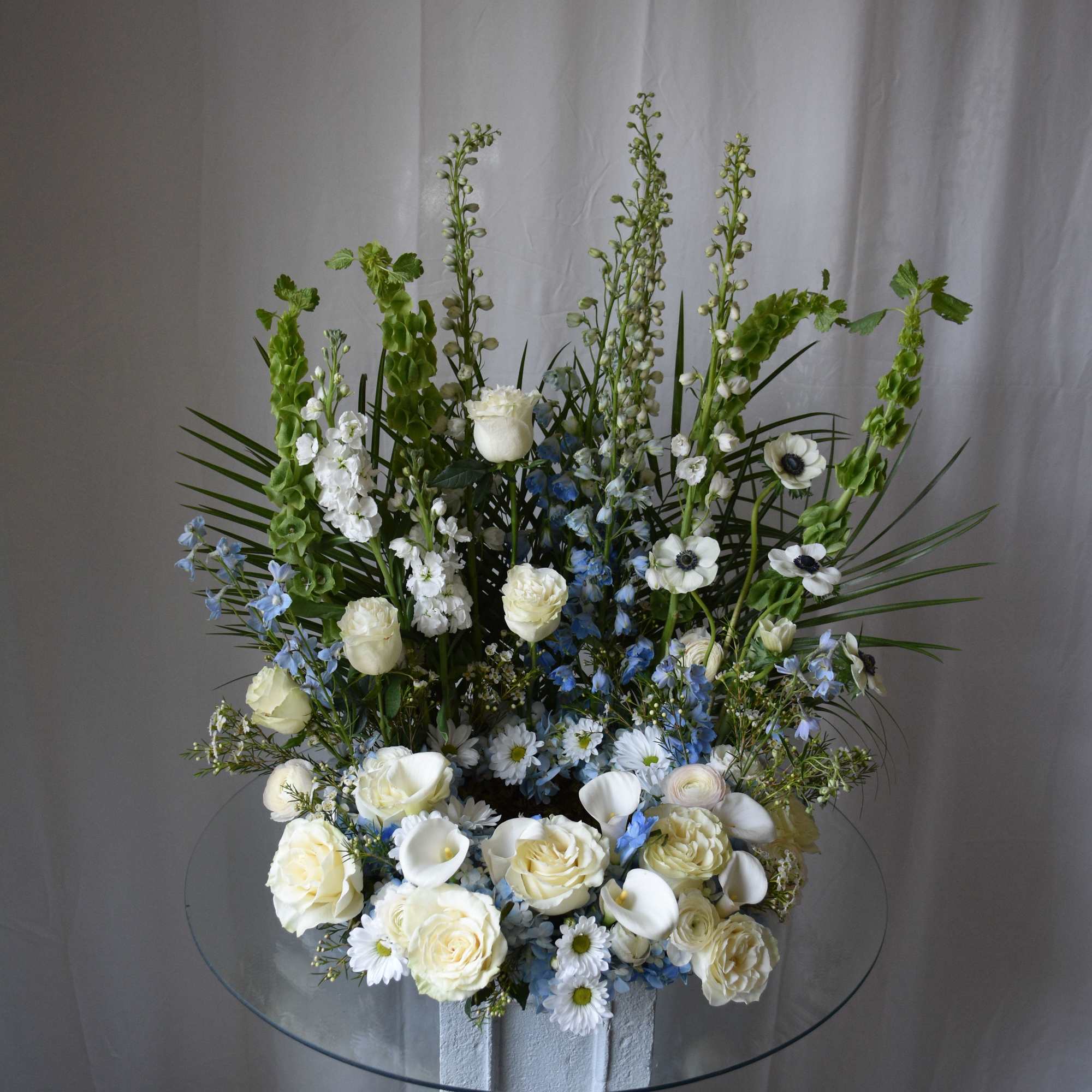 Tall white and blue floral arrangement with roses, calla lilies, and bells of Ireland on a glass-topped pedestal