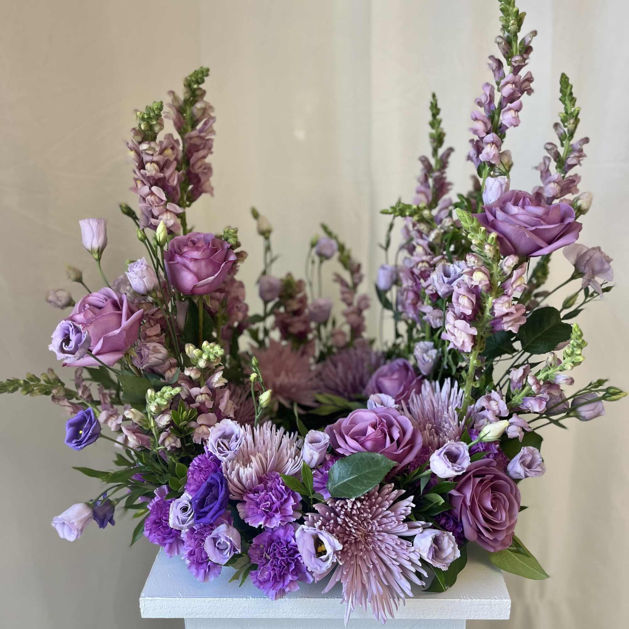 Lavender and purple floral arrangement with roses, snapdragons, and mums displayed on a white pedestal
