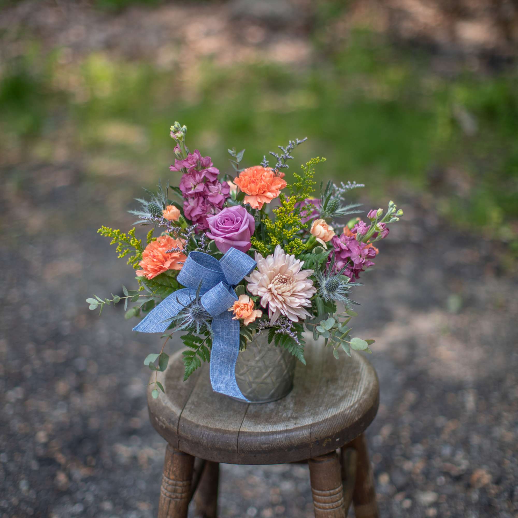 Mixed orange and purple flower arrangement with blue bow in a glass vase on a rustic wooden stool outdoors.