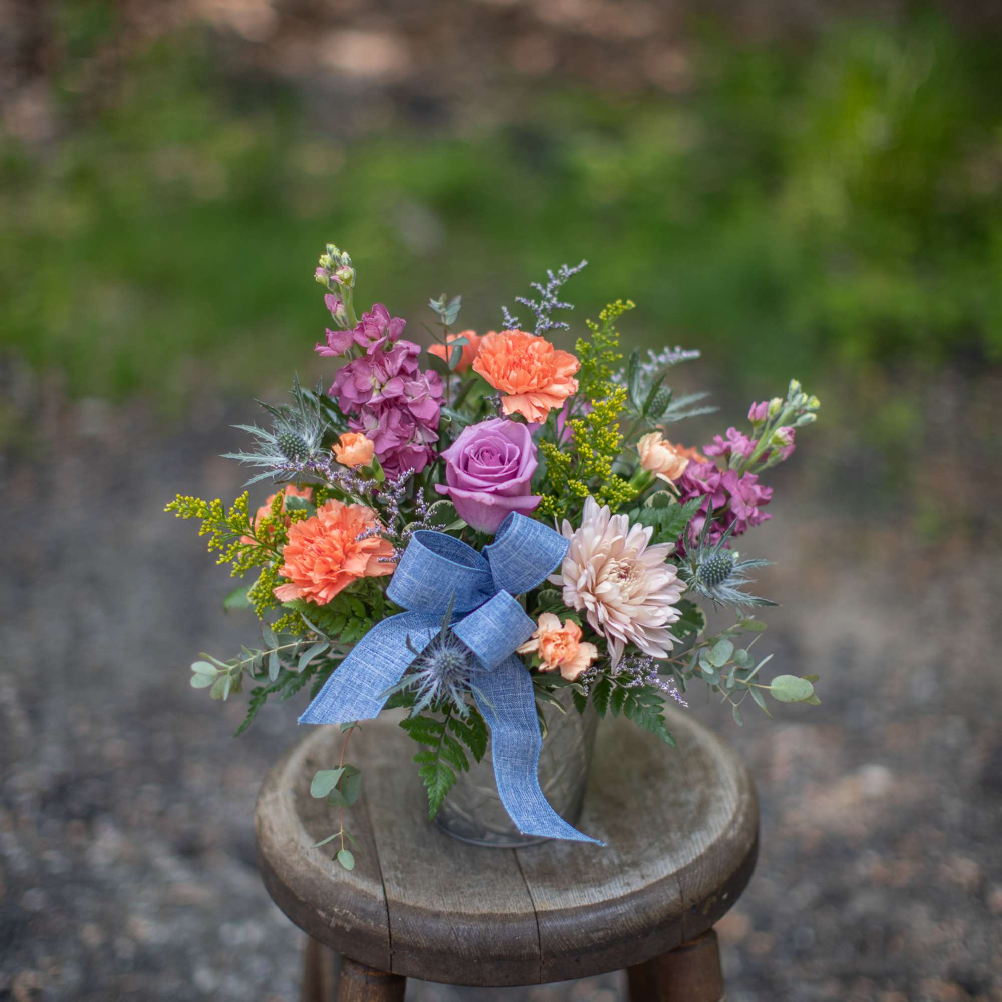 Compact arrangement of orange carnations and purple blooms in a small vase with a blue bow