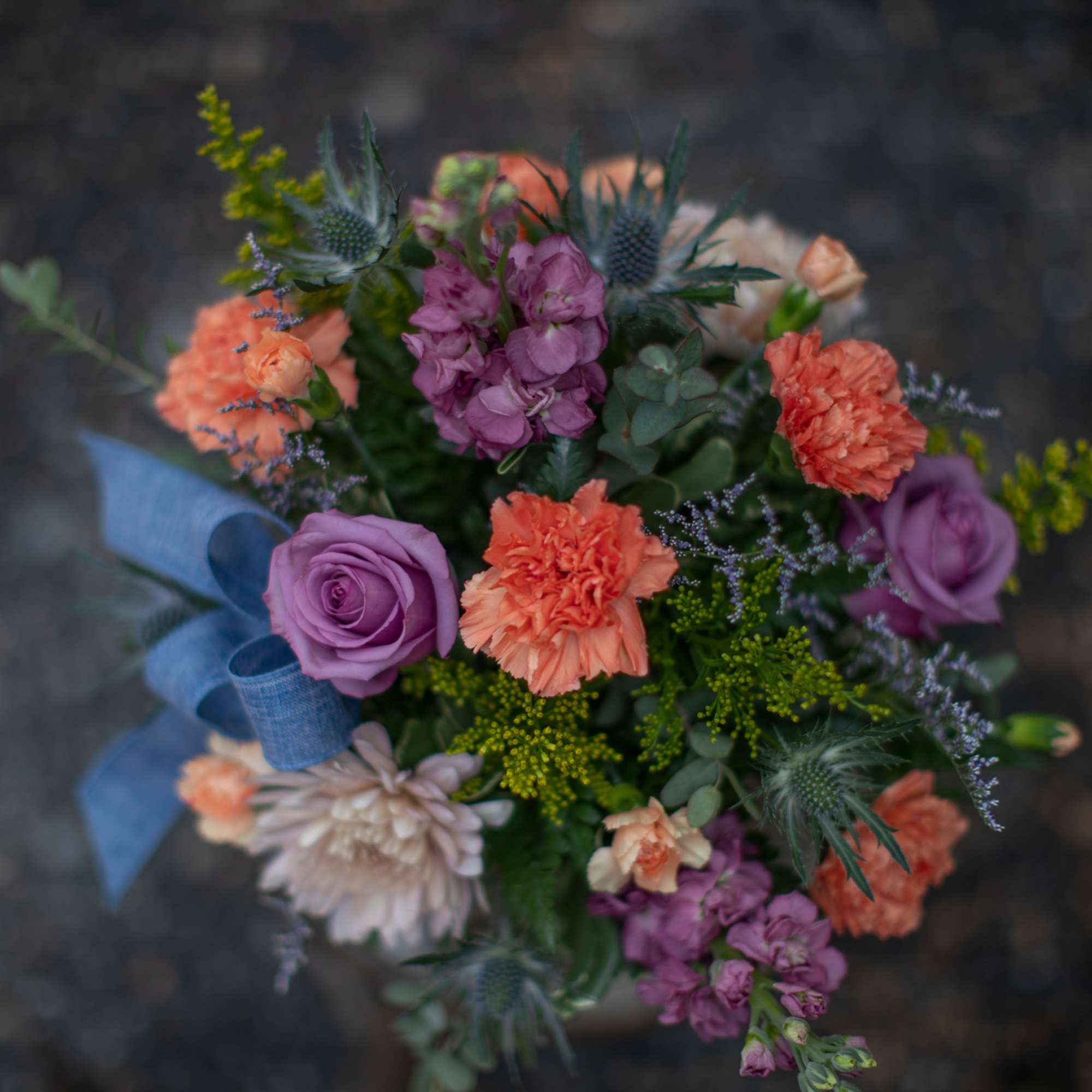 Round bouquet of lavender roses and orange carnations with accent flowers and a blue ribbon bow.