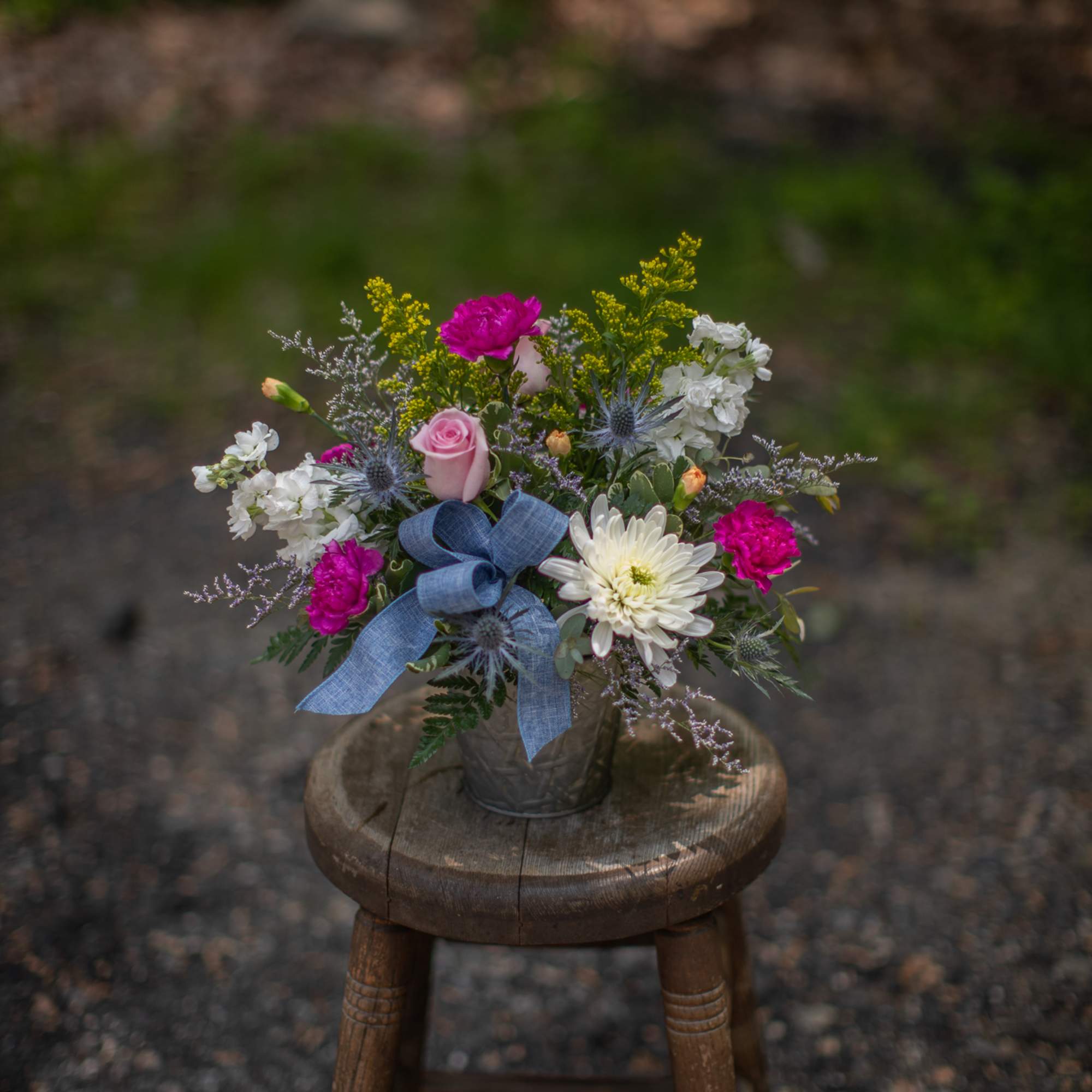 Mixed bouquet of pink roses, bright pink blooms, and white flowers in a metal pot with a blue bow.