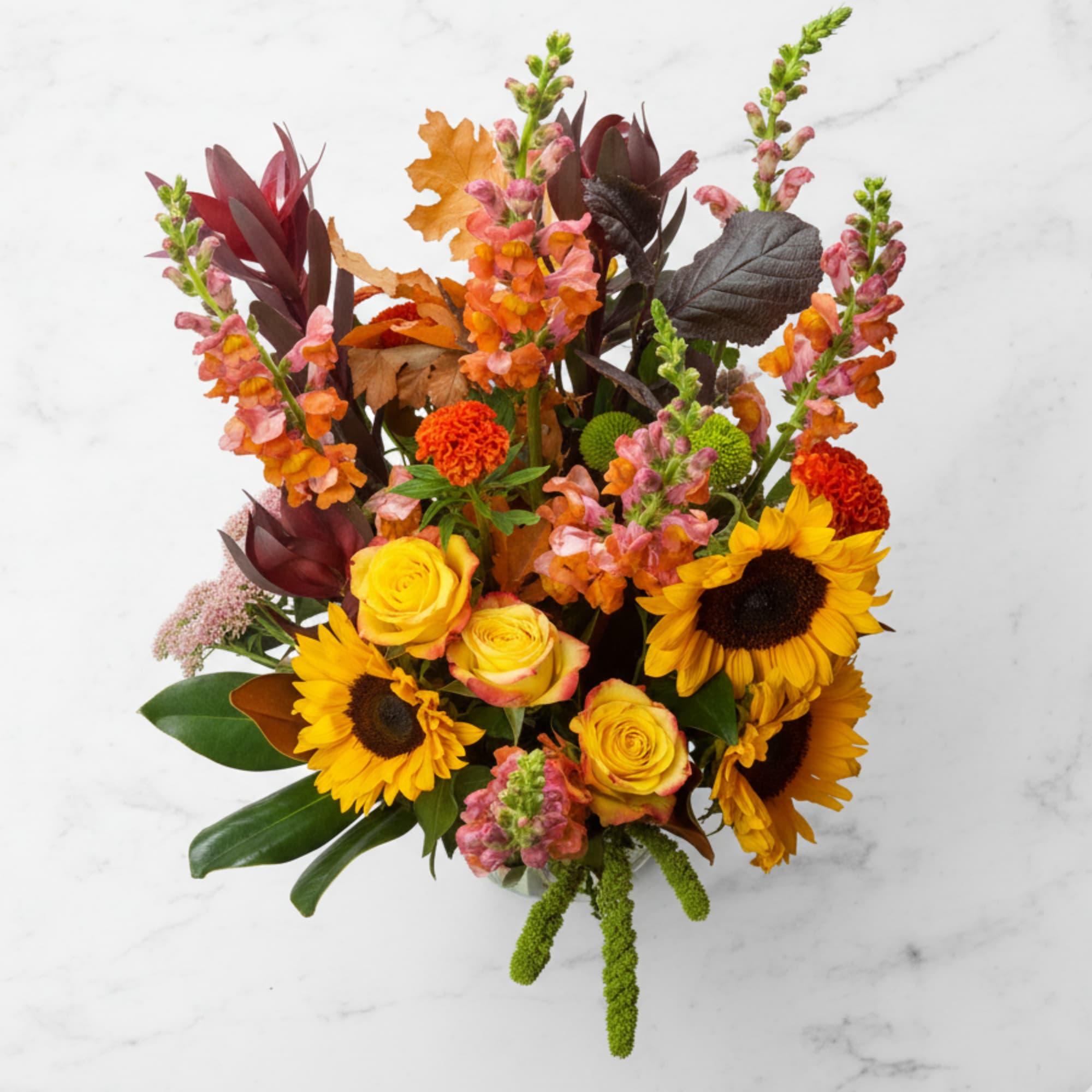 Autumn-toned arrangement with sunflowers, yellow roses, and orange blooms in a clear glass vase.