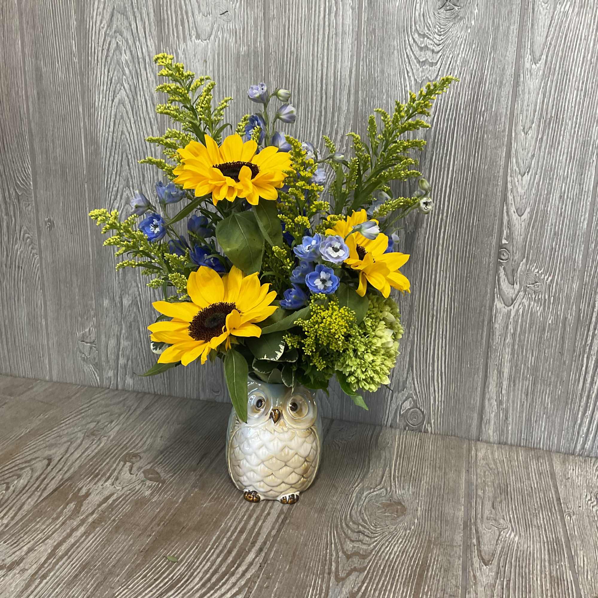 Yellow sunflowers and blue blooms arranged in a ceramic owl vase on a wood surface