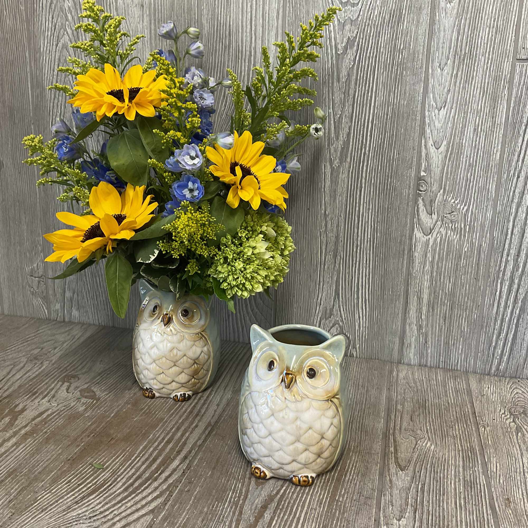 Yellow sunflowers and blue flowers in a ceramic owl vase beside a matching empty owl container.