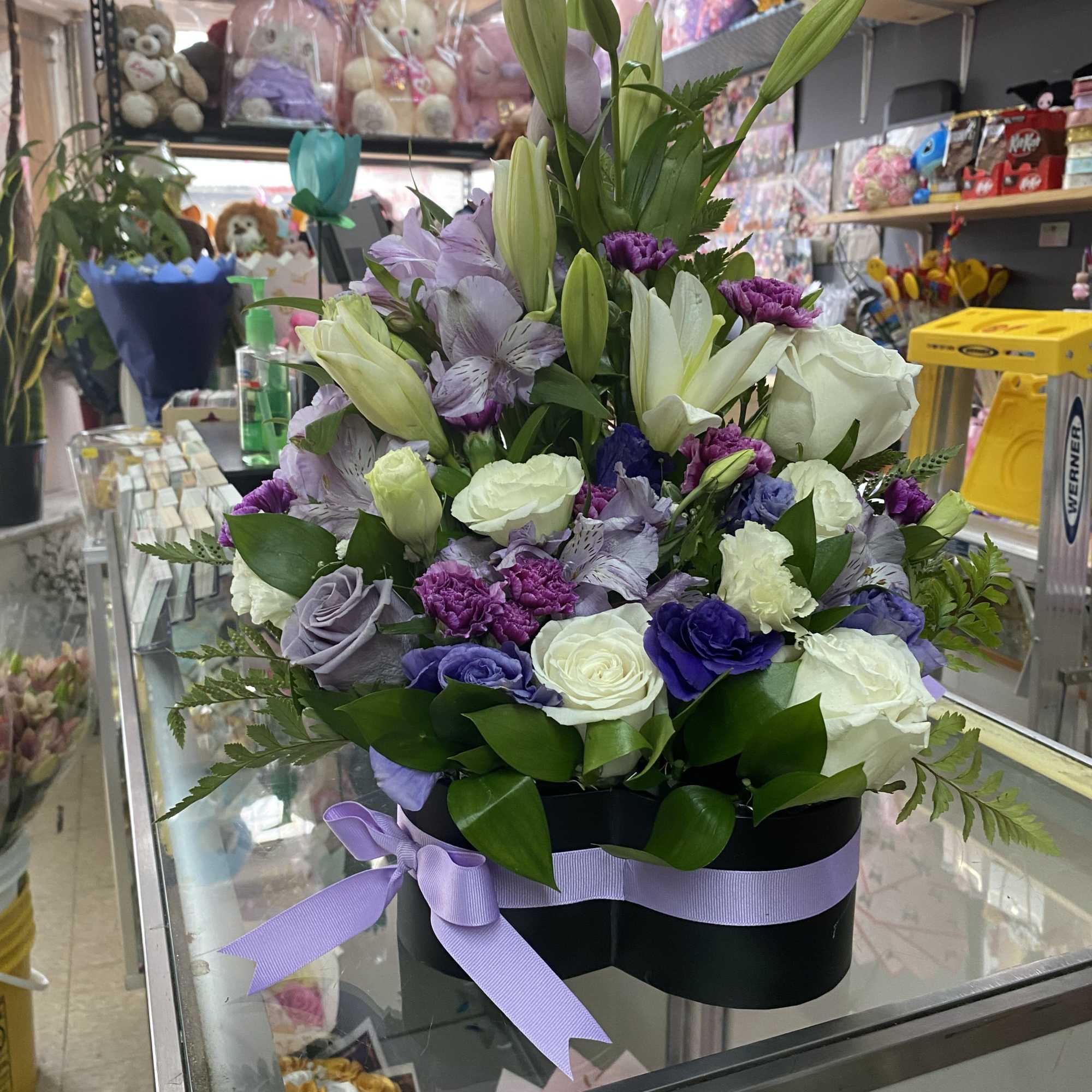 Heart box arrangement of white lilies, roses, and purple flowers with a lavender ribbon bow
