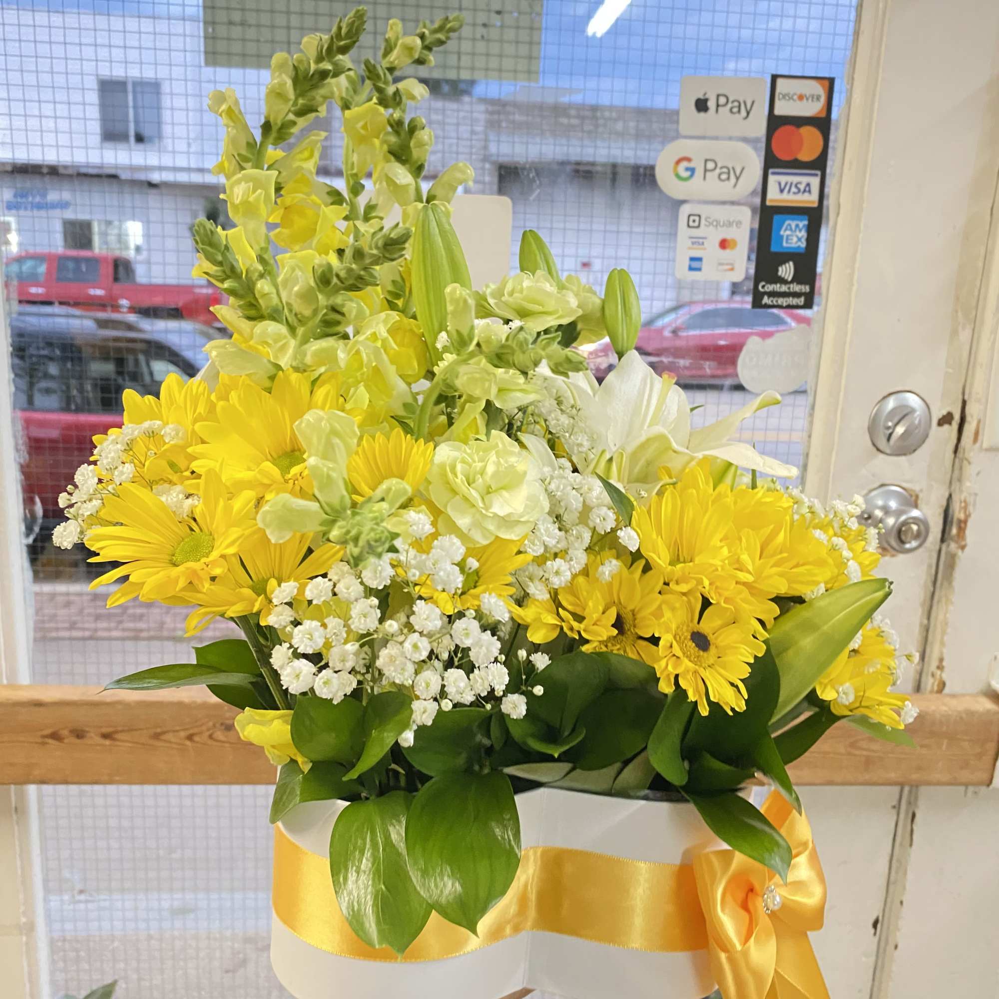 Yellow and white flower arrangement with daisies, lilies, and snapdragons in a white box tied with a yellow ribbon.