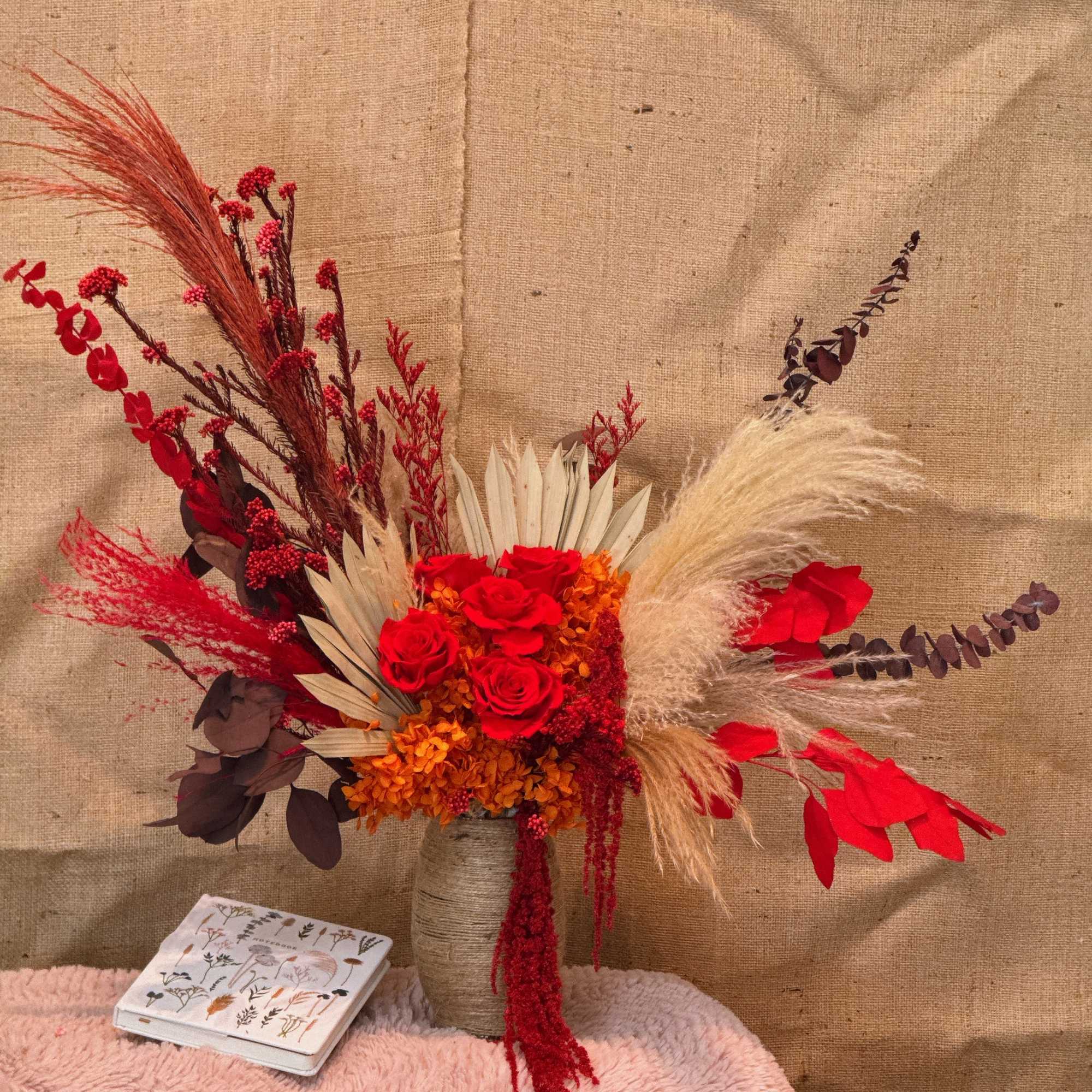 Asymmetrical arrangement of red roses, orange blooms, and dried pampas in a tall twine-wrapped vase.