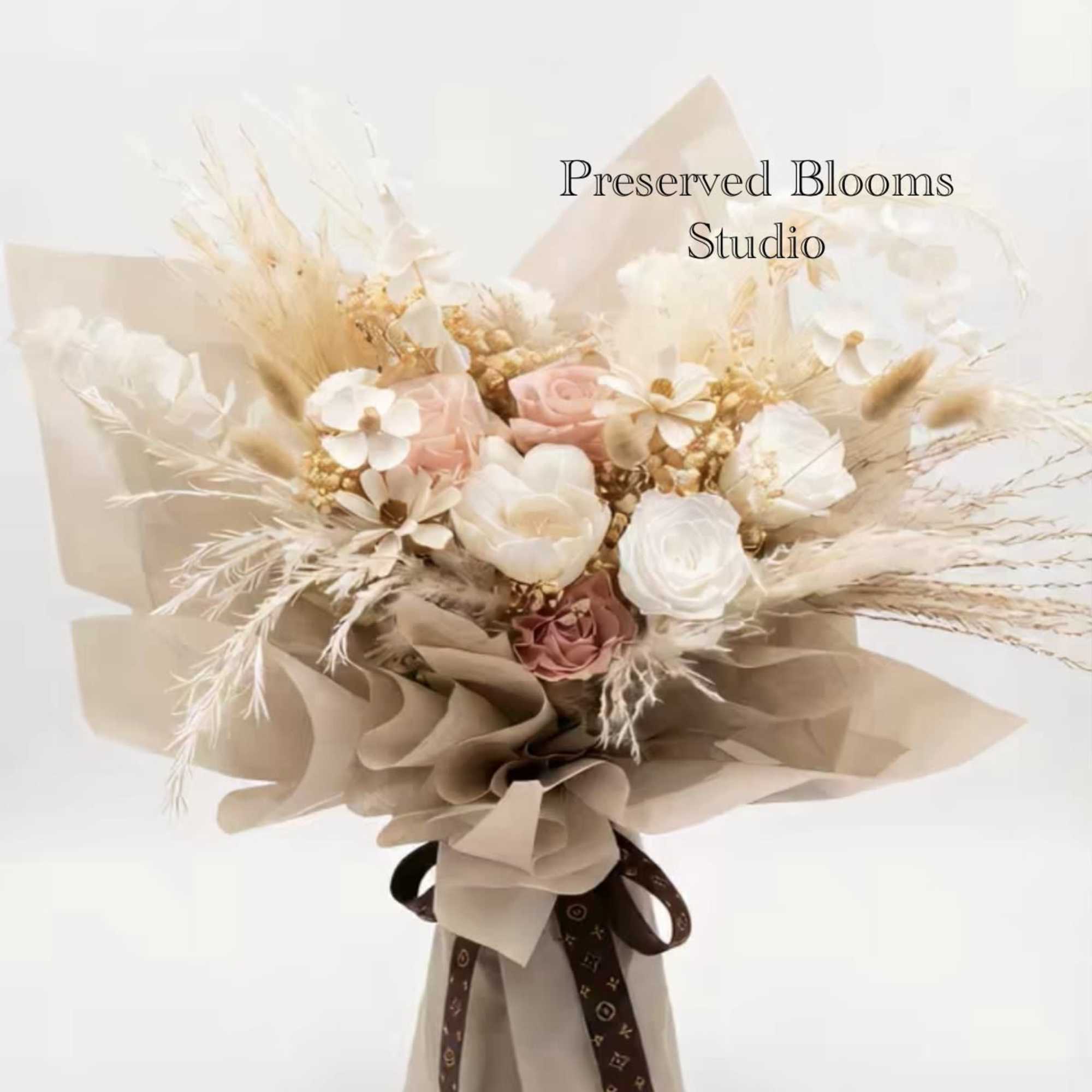 Bouquet of preserved white and blush flowers with dried grasses wrapped in tan paper and tied with a brown ribbon