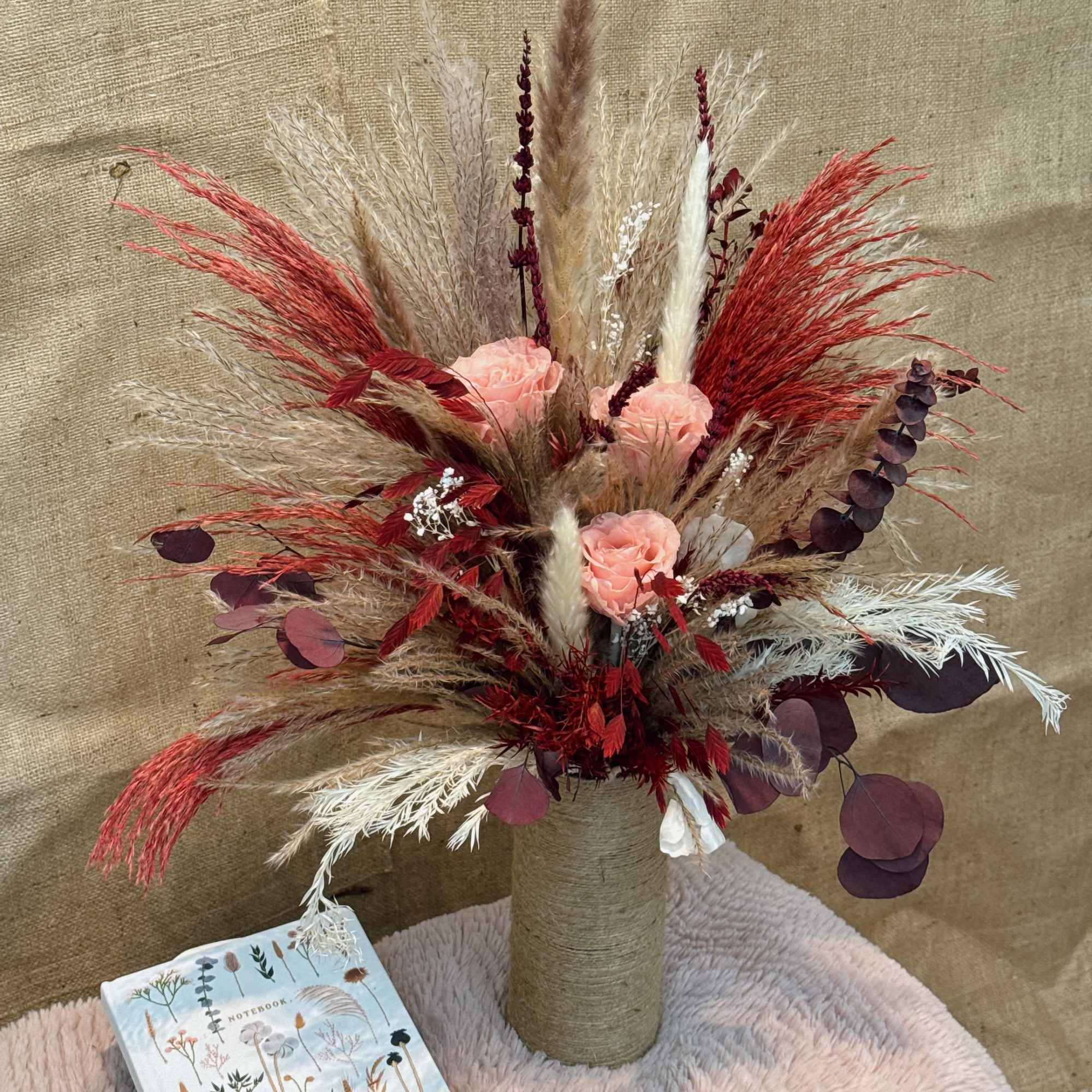Tall dried grass arrangement with peach roses in a twine-wrapped vase beside a floral notebook