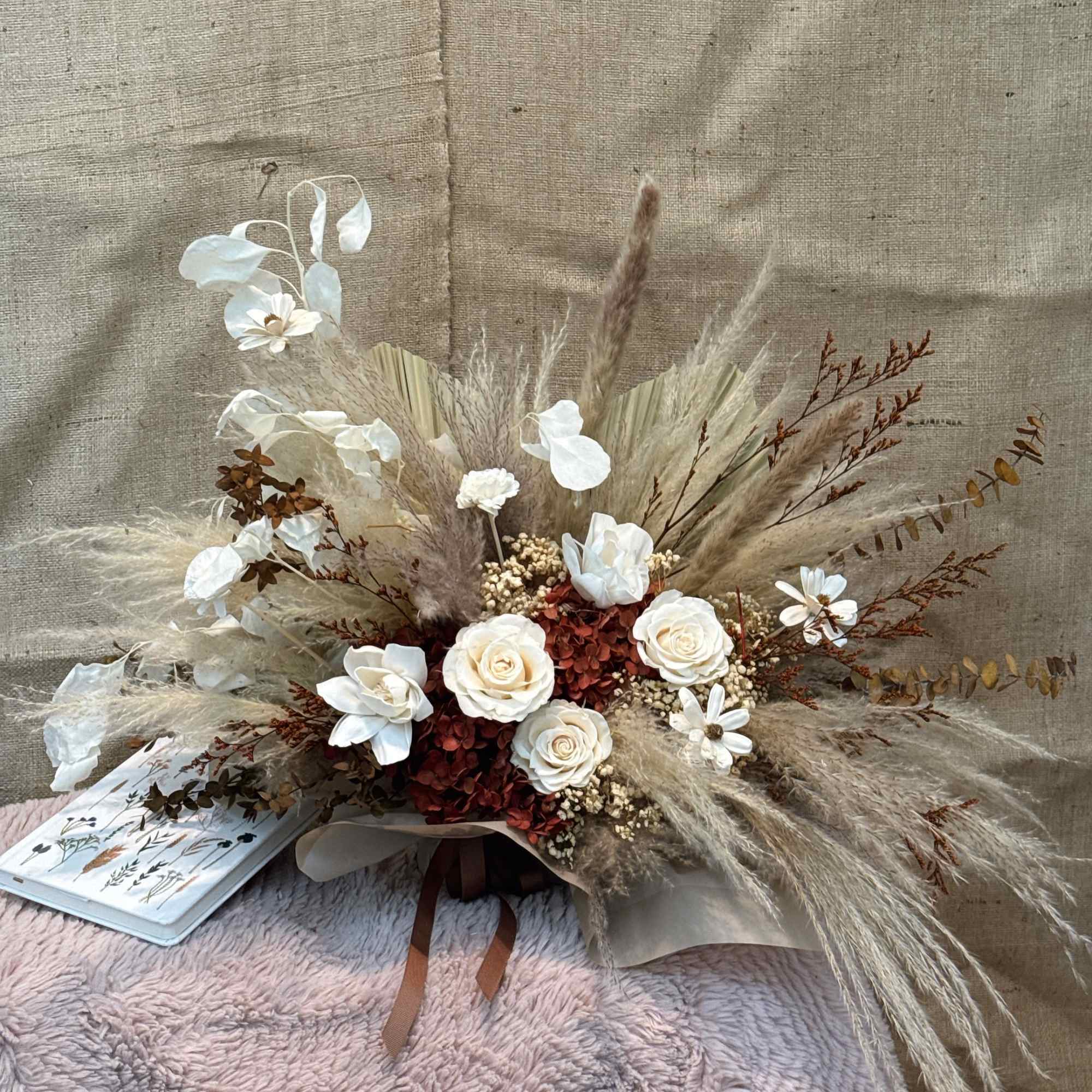 Low dried arrangement with white roses, rust blooms, and pampas grass in neutral wrapping on a textured throw.