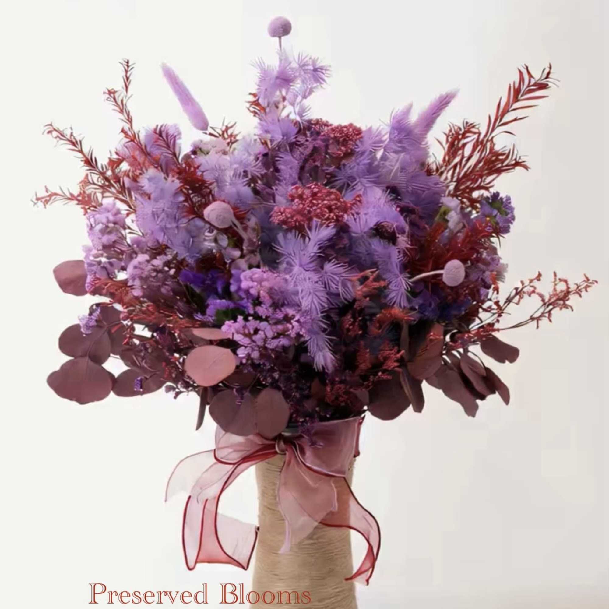 Tall bouquet of lavender and burgundy preserved flowers in a rope-wrapped vase with a pink ribbon bow