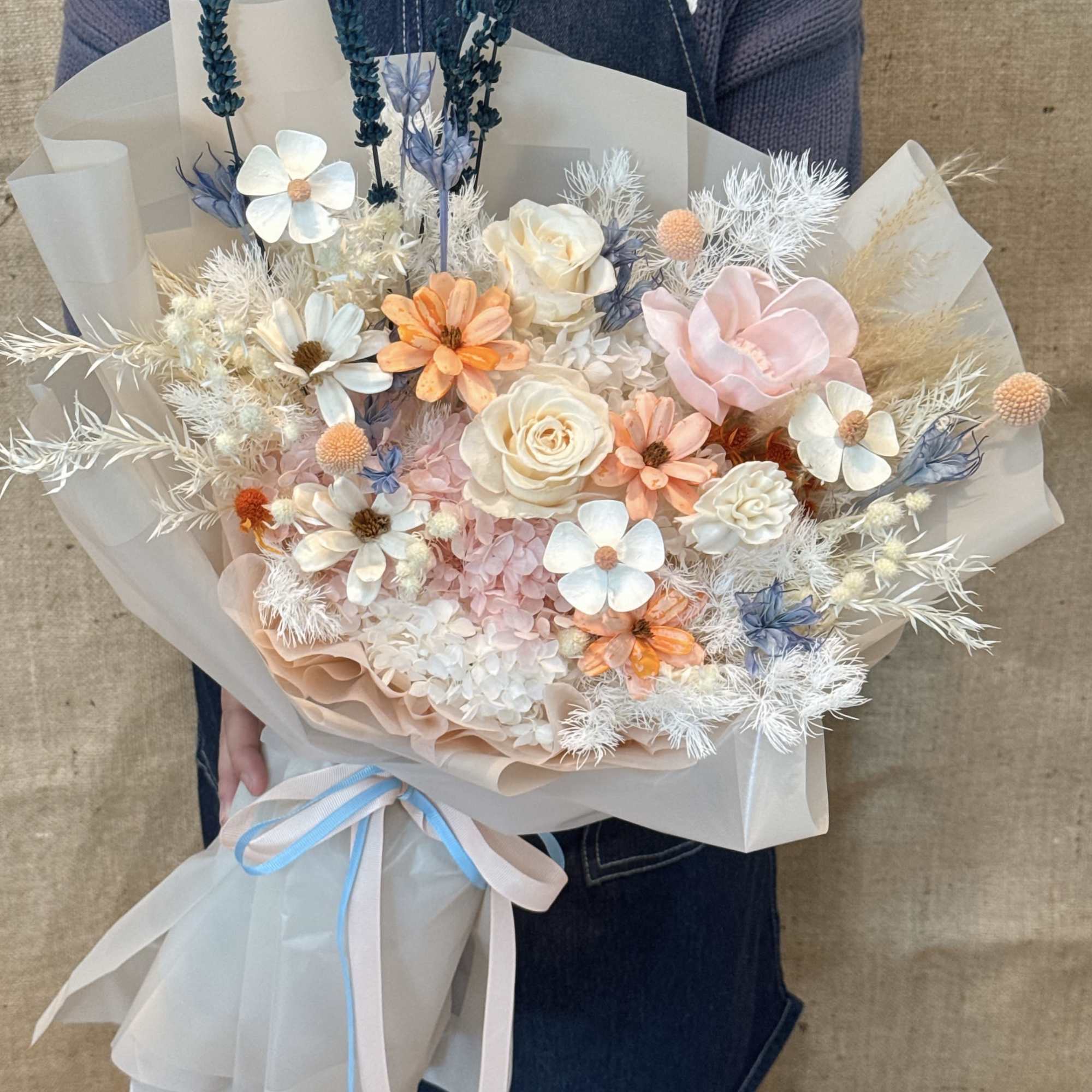 Large hand-tied bouquet of peach, white, and blush flowers with blue accents wrapped in soft beige paper
