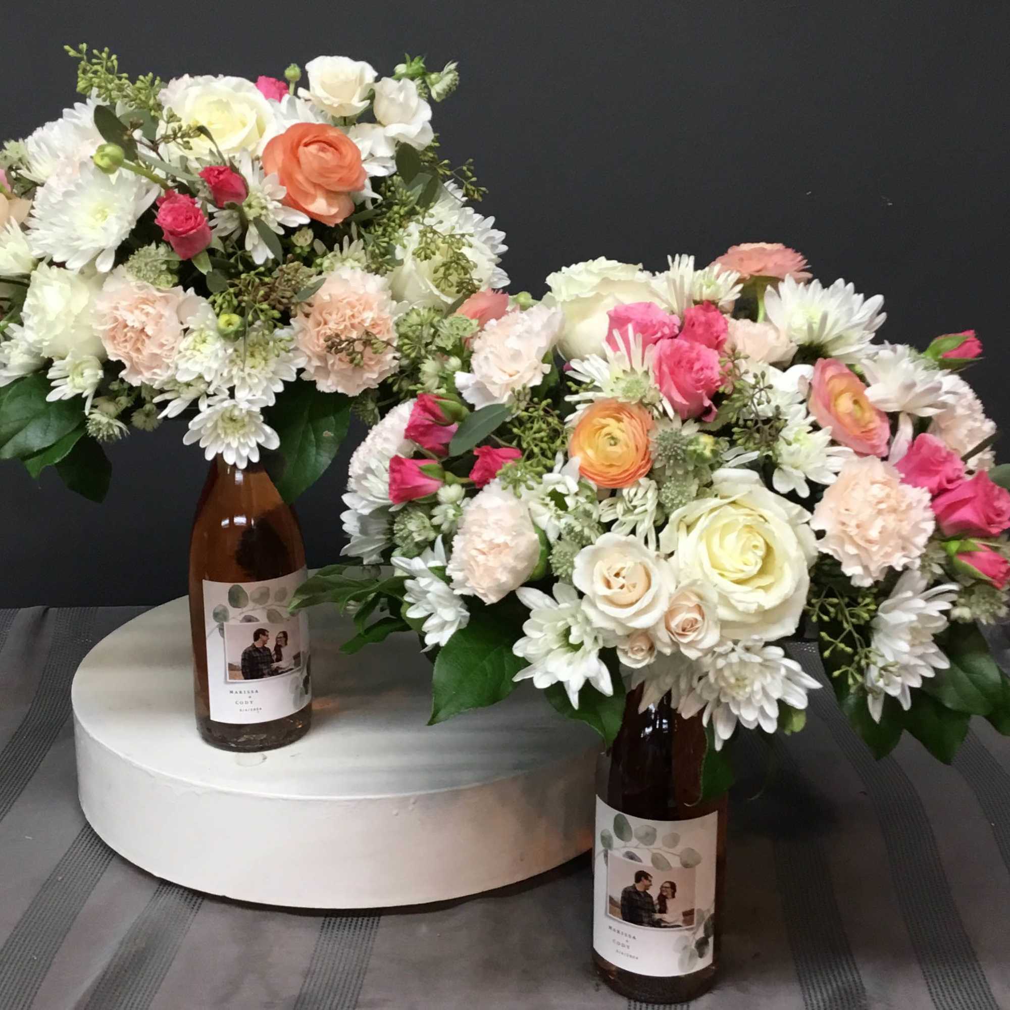 Two wedding centerpieces of white and pastel roses, ranunculus, carnations, and mums arranged in labeled brown bottles