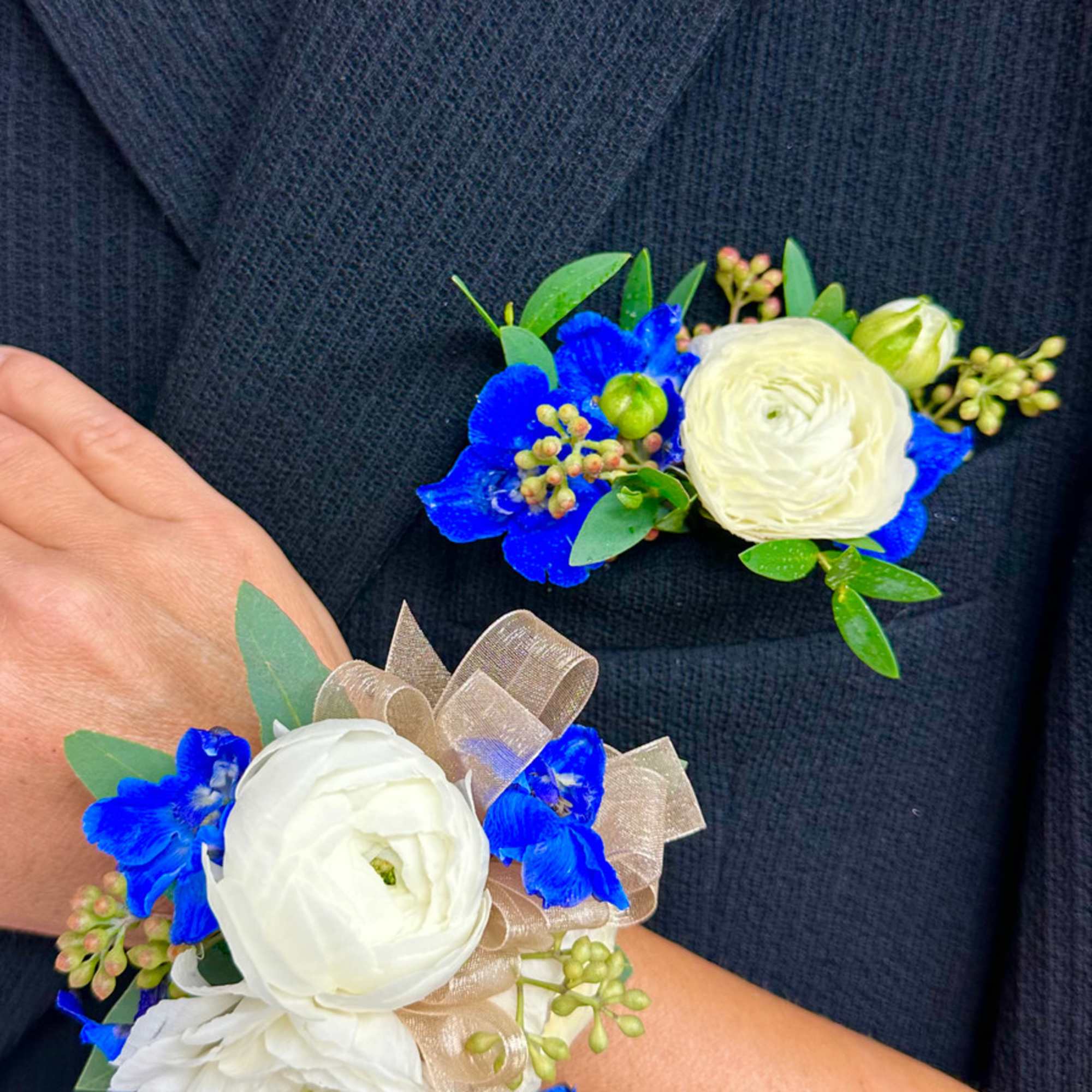 White and blue floral wrist corsage and matching boutonniere on a dark suit jacket