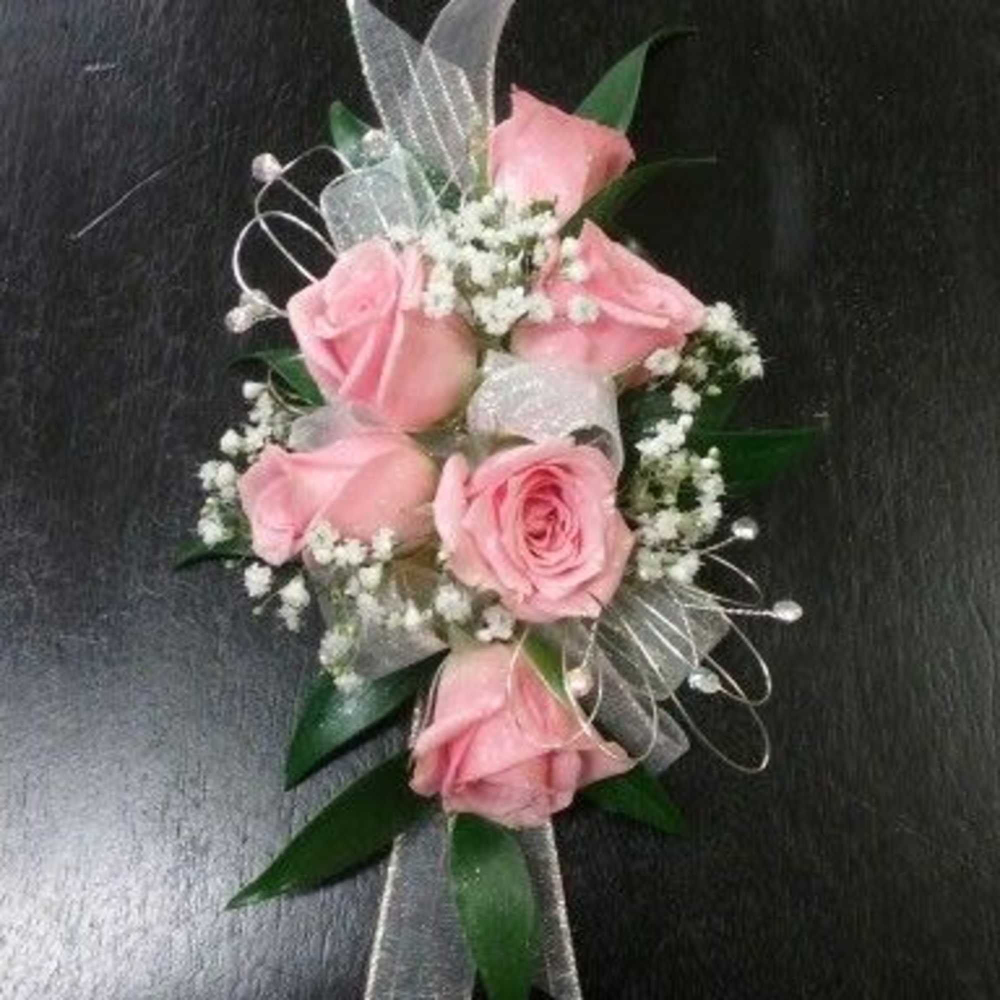 Pink rose wrist corsage with white filler flowers and sheer ribbon on a dark surface