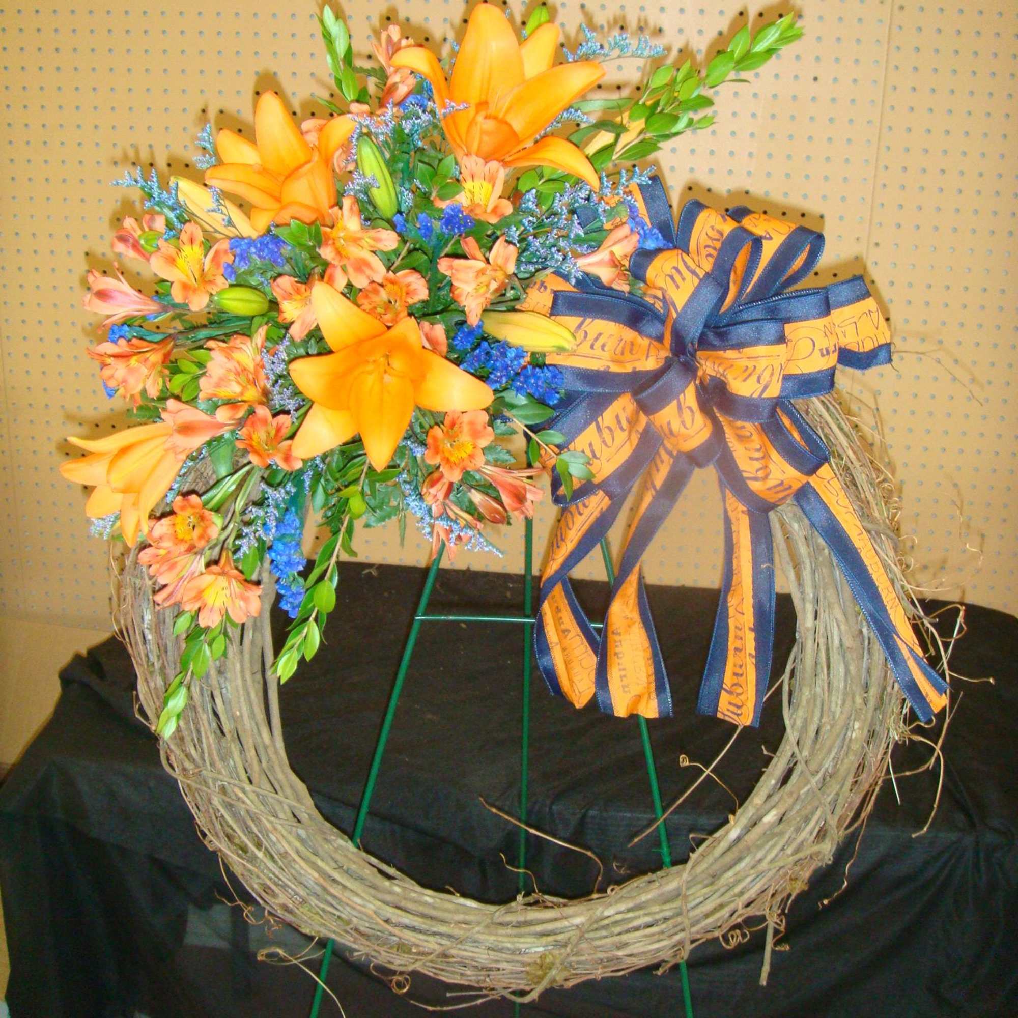 Grapevine wreath on an easel with orange lilies, orange blooms, blue accents, and a large orange and navy bow.