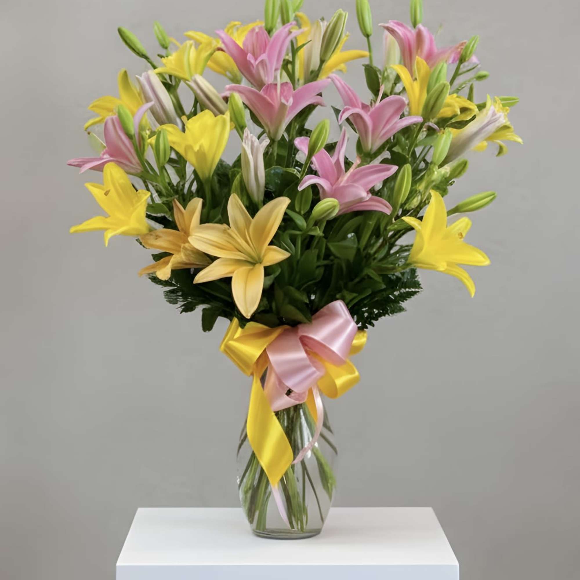 Tall clear vase of pink and yellow lilies with buds, tied with a pink and yellow ribbon bow.
