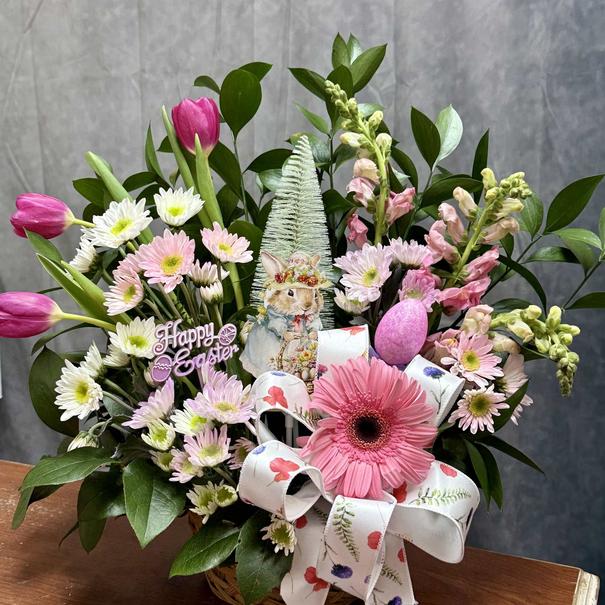 Easter basket with pink tulips, daisies, gerbera daisy and snapdragons with bunny and egg decorations.