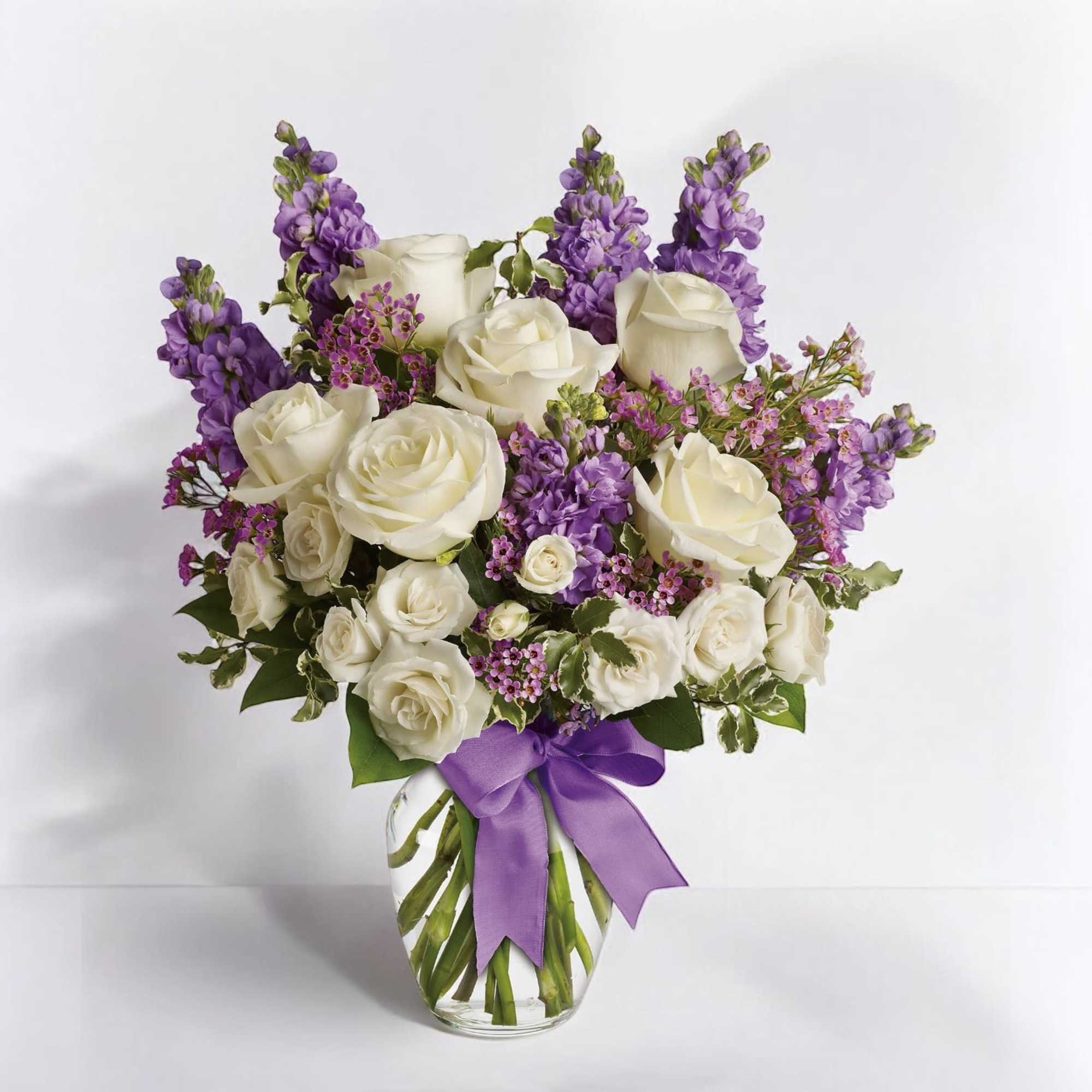White roses and purple flowers arranged in a clear glass vase with a purple ribbon