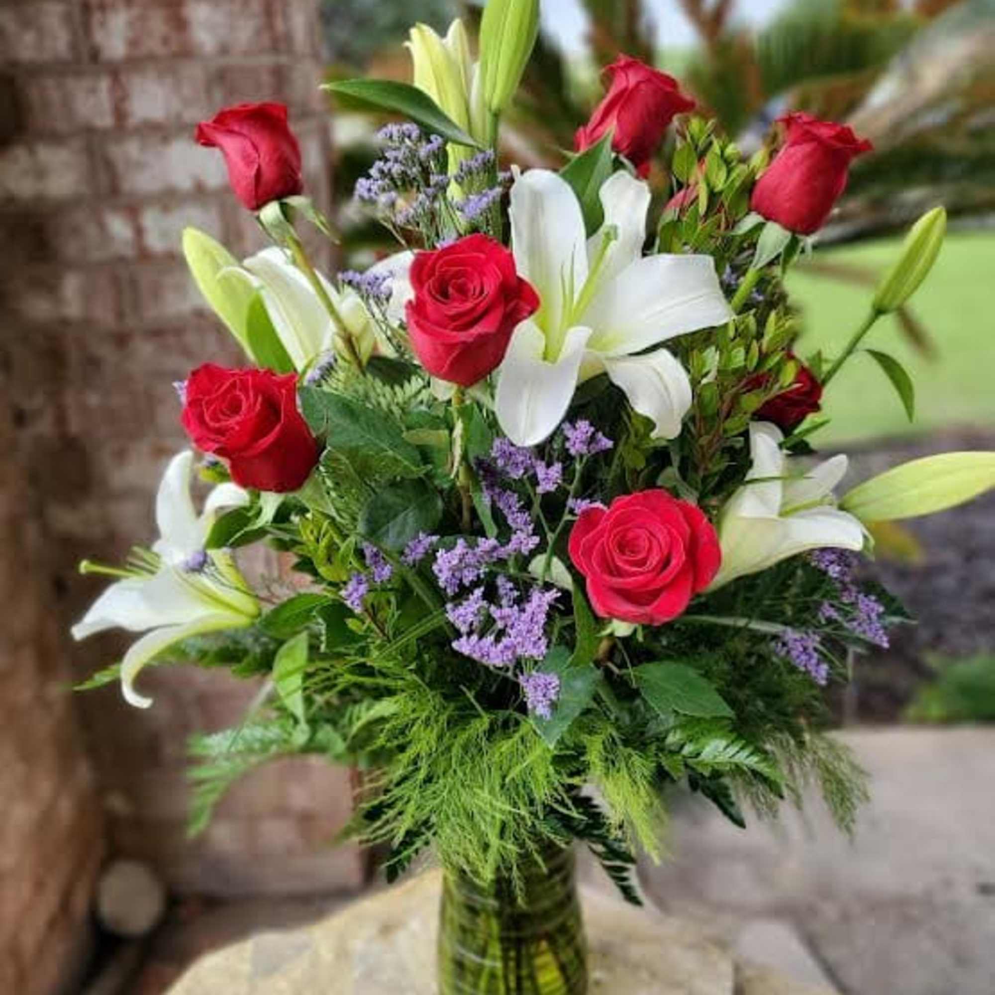 Tall arrangement of red roses, white lilies, and purple flowers in a green glass vase